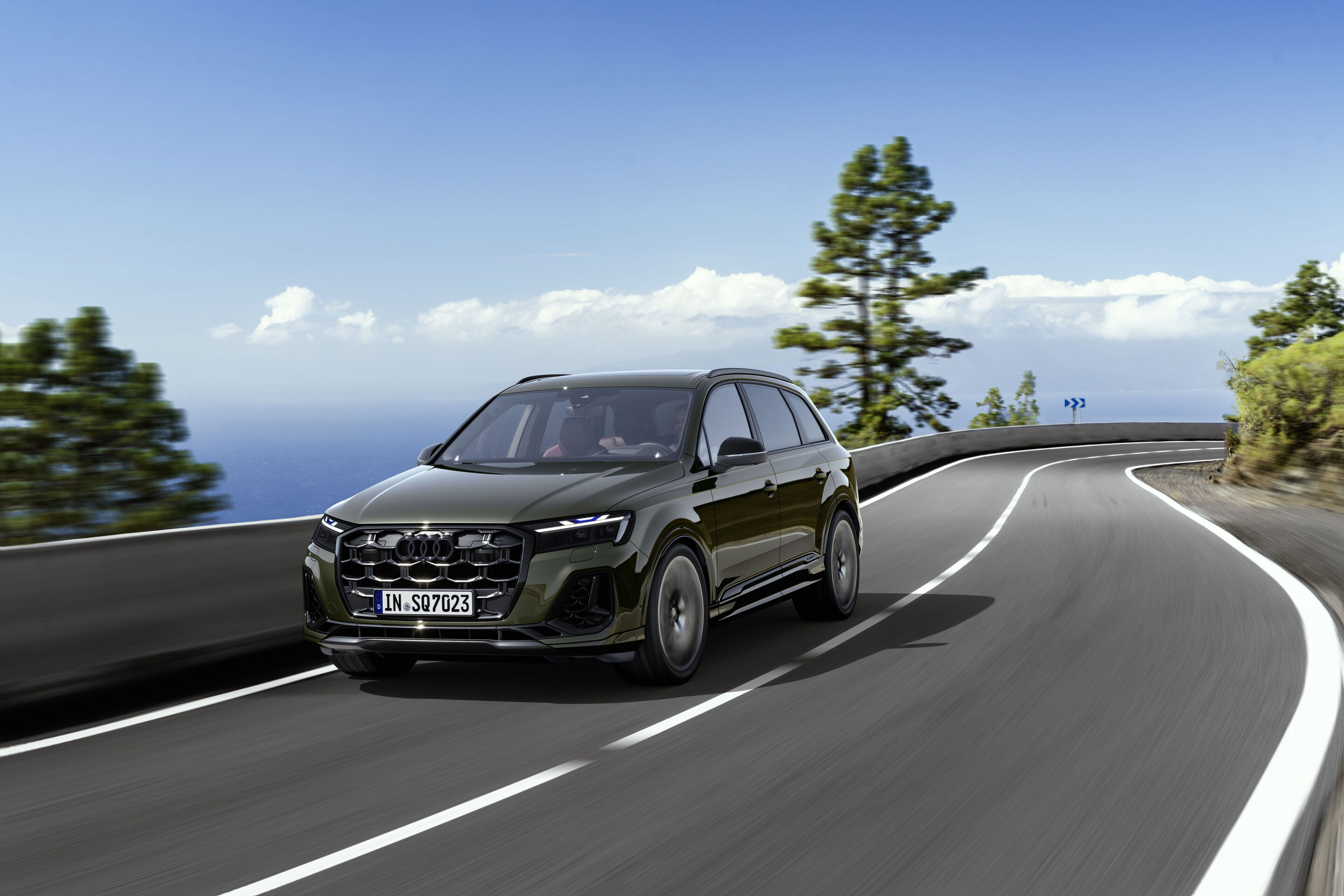 Audi SQ7 TFSI (until 2026) - An Audi SQ7 in Oak green pearl effect drives along a coastal road lined with pines. The sea can be seen in the background.