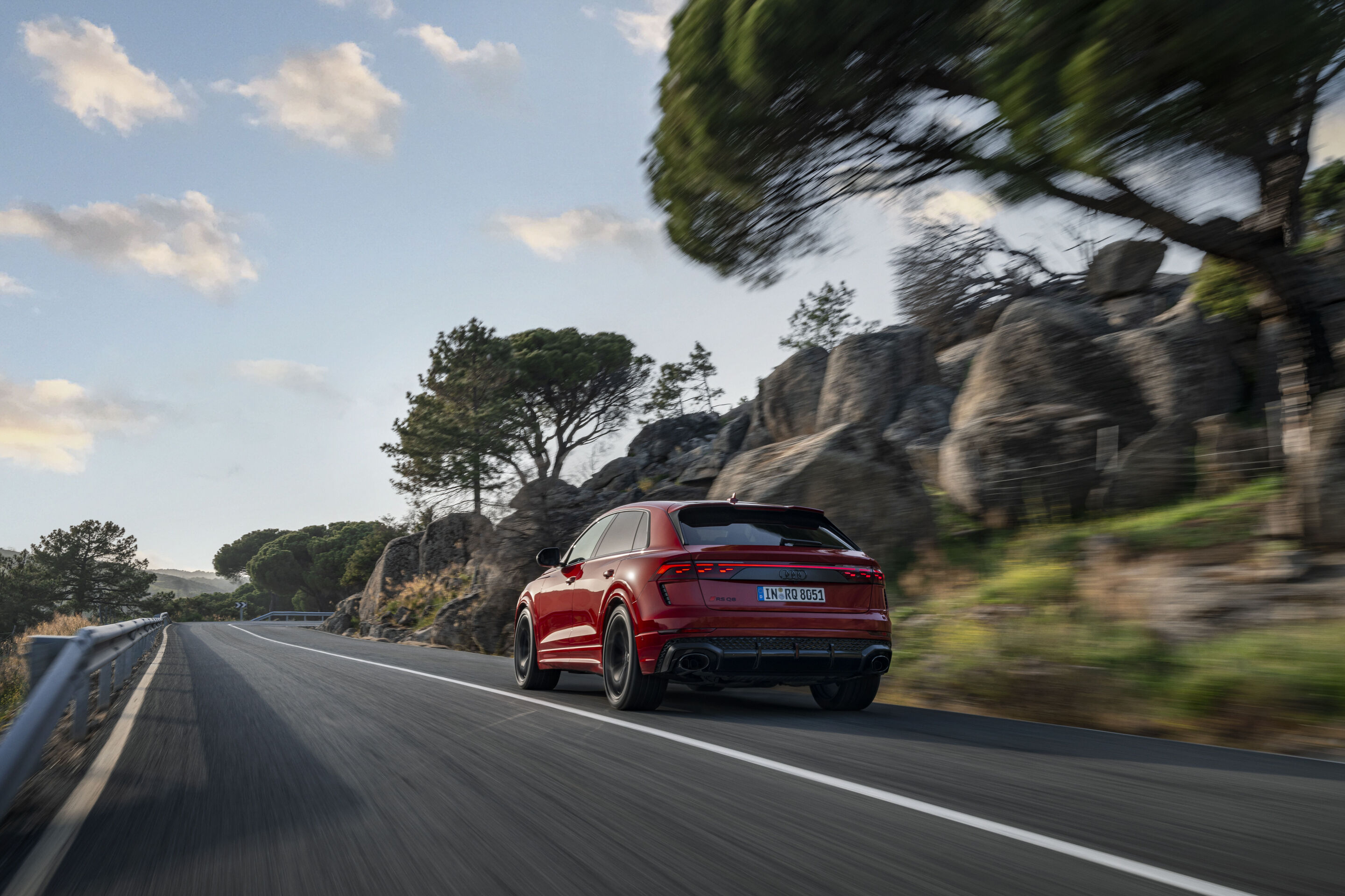 Audi RS Q8 performance - An Audi RS Q8 in bright Chili red metallic drives along a winding country road lined with rock formations and rugged vegetation.