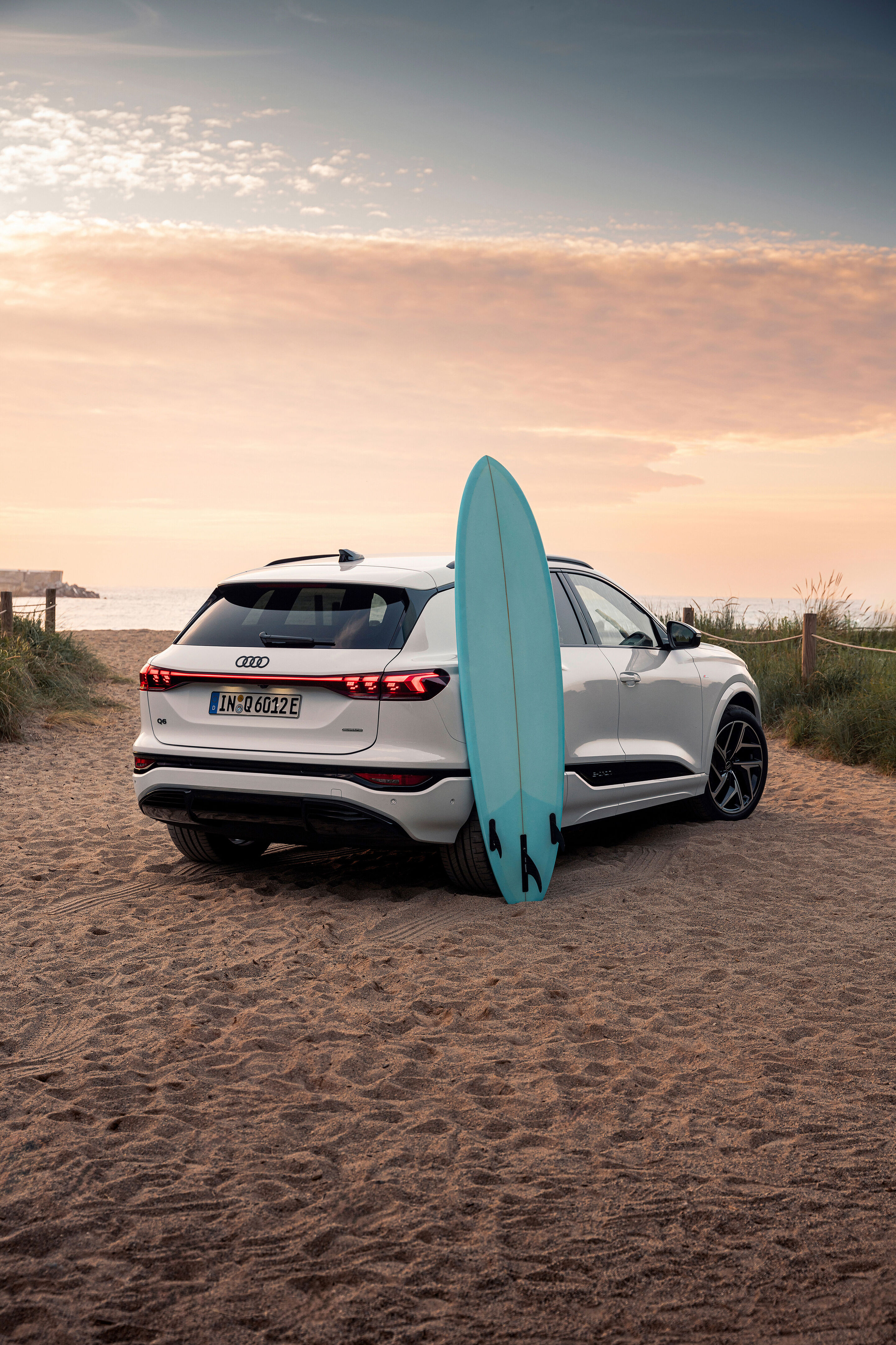 Audi Q6 e-tron quattro - Ein Audi Q6 e-tron in Gletscherweiß Metallic steht am Strand zwischen Dünengräsern. Am Heck lehnt ein Surfboard.