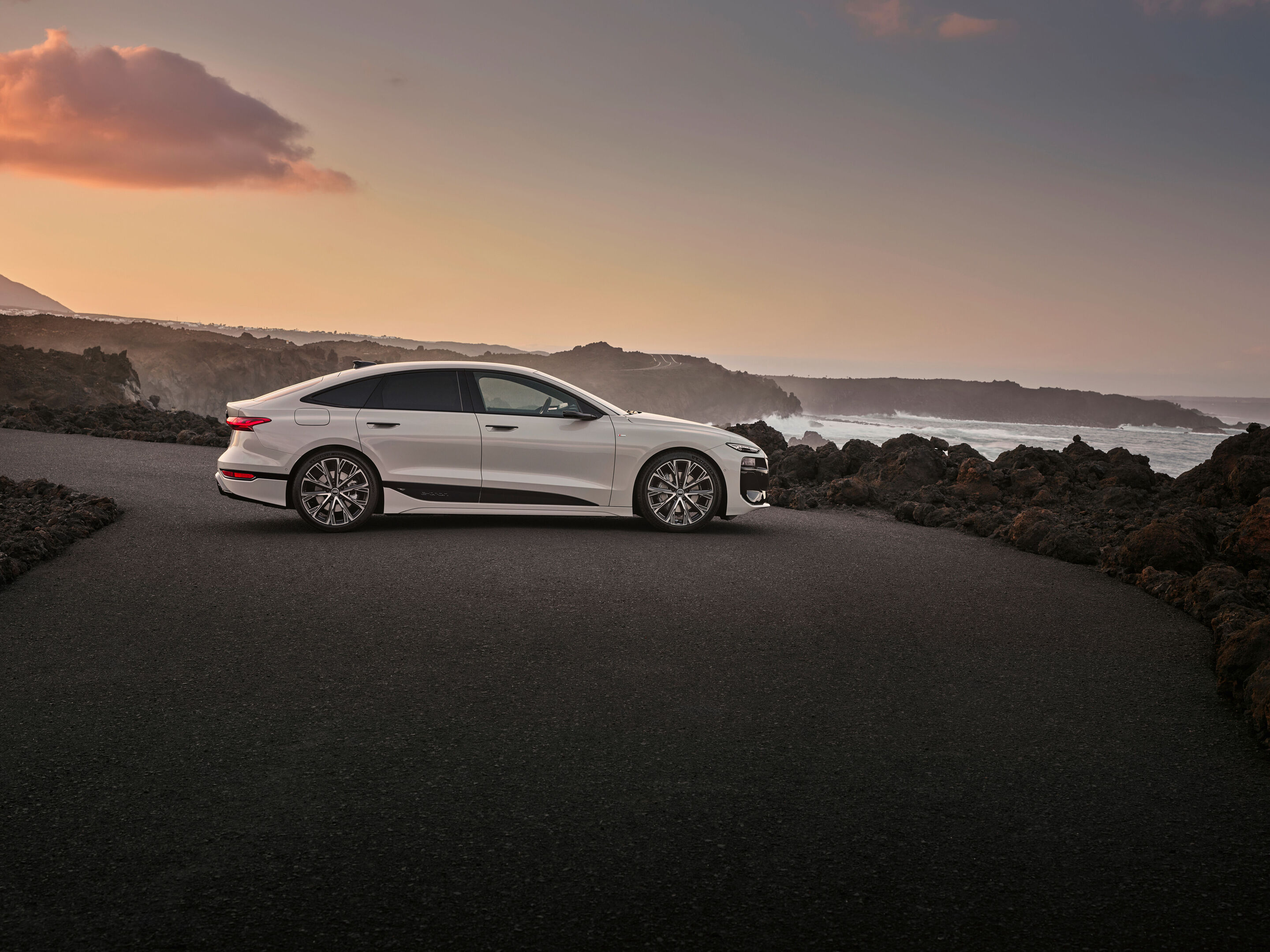 Audi A6 Sportback e-tron performance - Ein Audi A6 Sportback e-tron in Siambeige Metallic steht auf einem Parkplatz unmittelbar an einer Felsformation. Im Hintergrund bricht das Meer an der Küste.