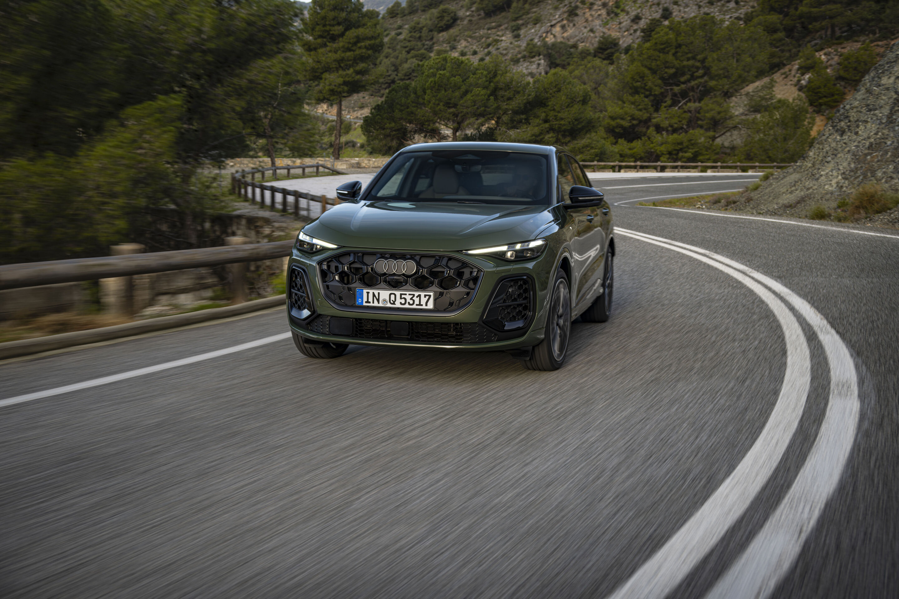 Audi Q5 Sportback - Ein dunkelgrüner SUV fährt auf einer kurvigen Bergstraße entlang, umgeben von Bäumen und Felsen. Das Fahrzeug bewegt sich leicht nach links in die Kurve.