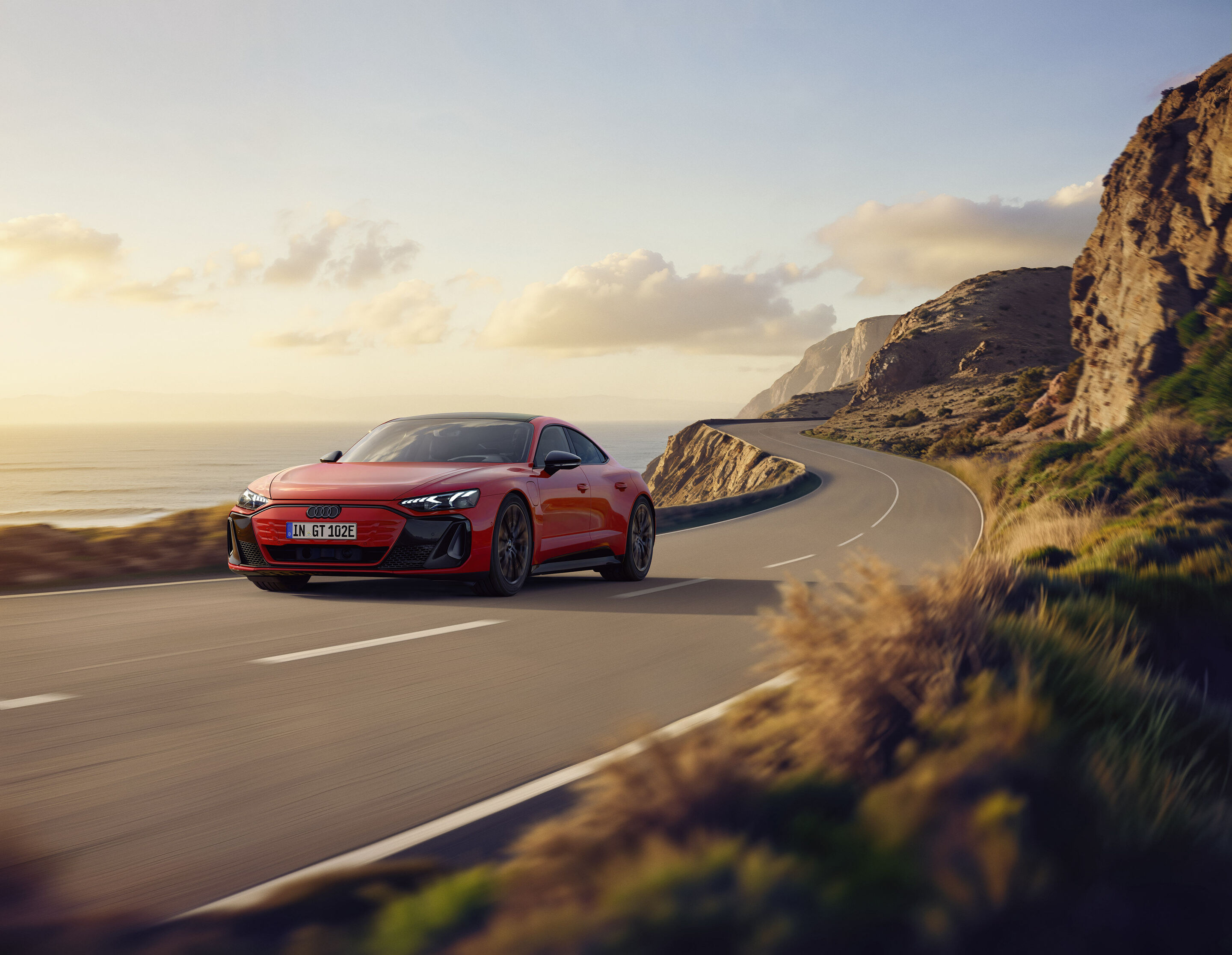 Einstieg in die Spitzenklasse: Audi e-tron GT quattro vereint emotionales Design, Performance und Komfort - Ein Audi e-tron GT quattro in Progressivrot Metallic fährt eine Küstenstraße entlang. Im Hintergrund befinden sich Berge und das Meer.