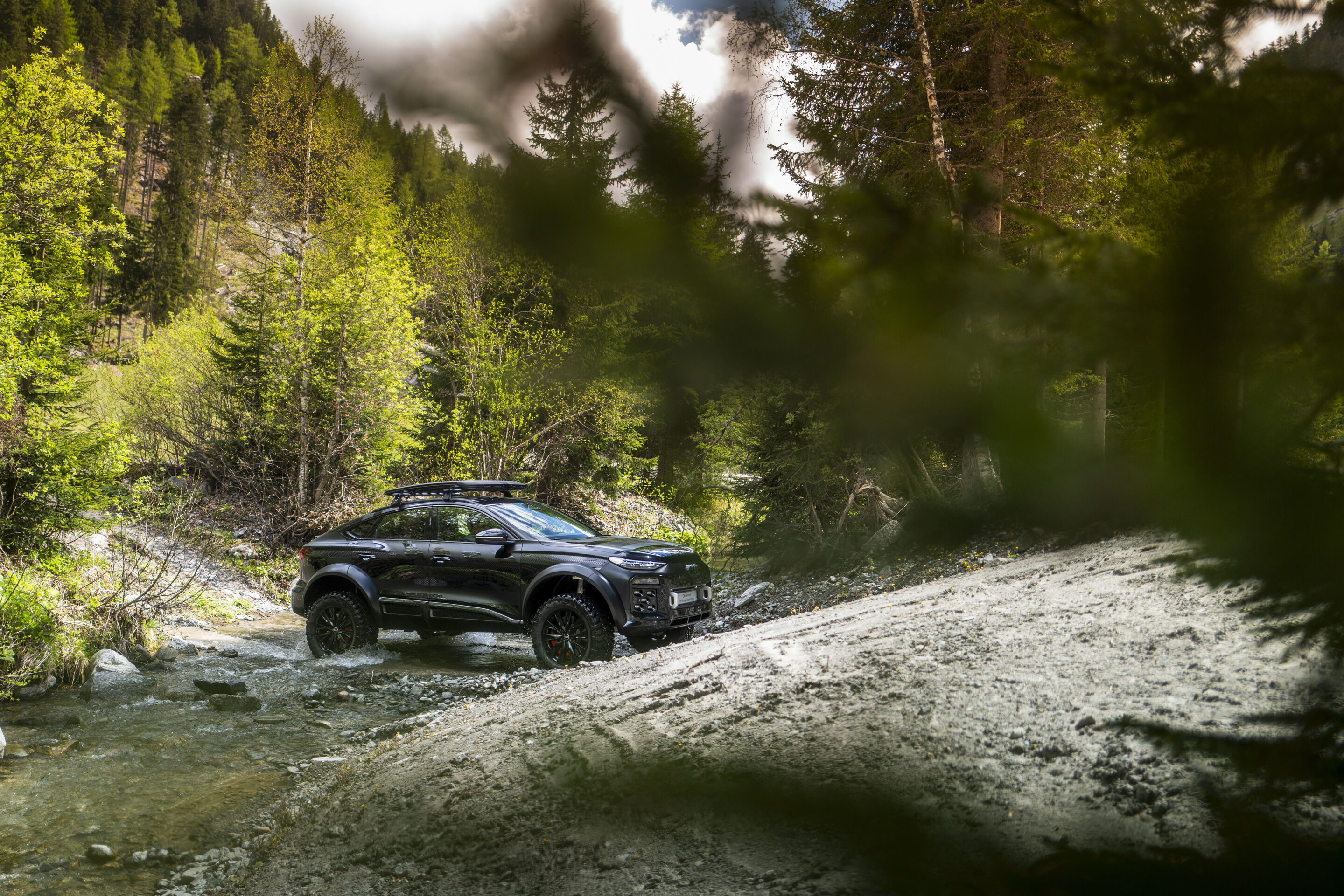 Audi Q6 e-tron offroad concept im Kaunertal - Der Audi Q6 e-tron offroad concept durchquert im österreichischen Kauntertal, umgeben von einem Mischwald, einen Fluss.
