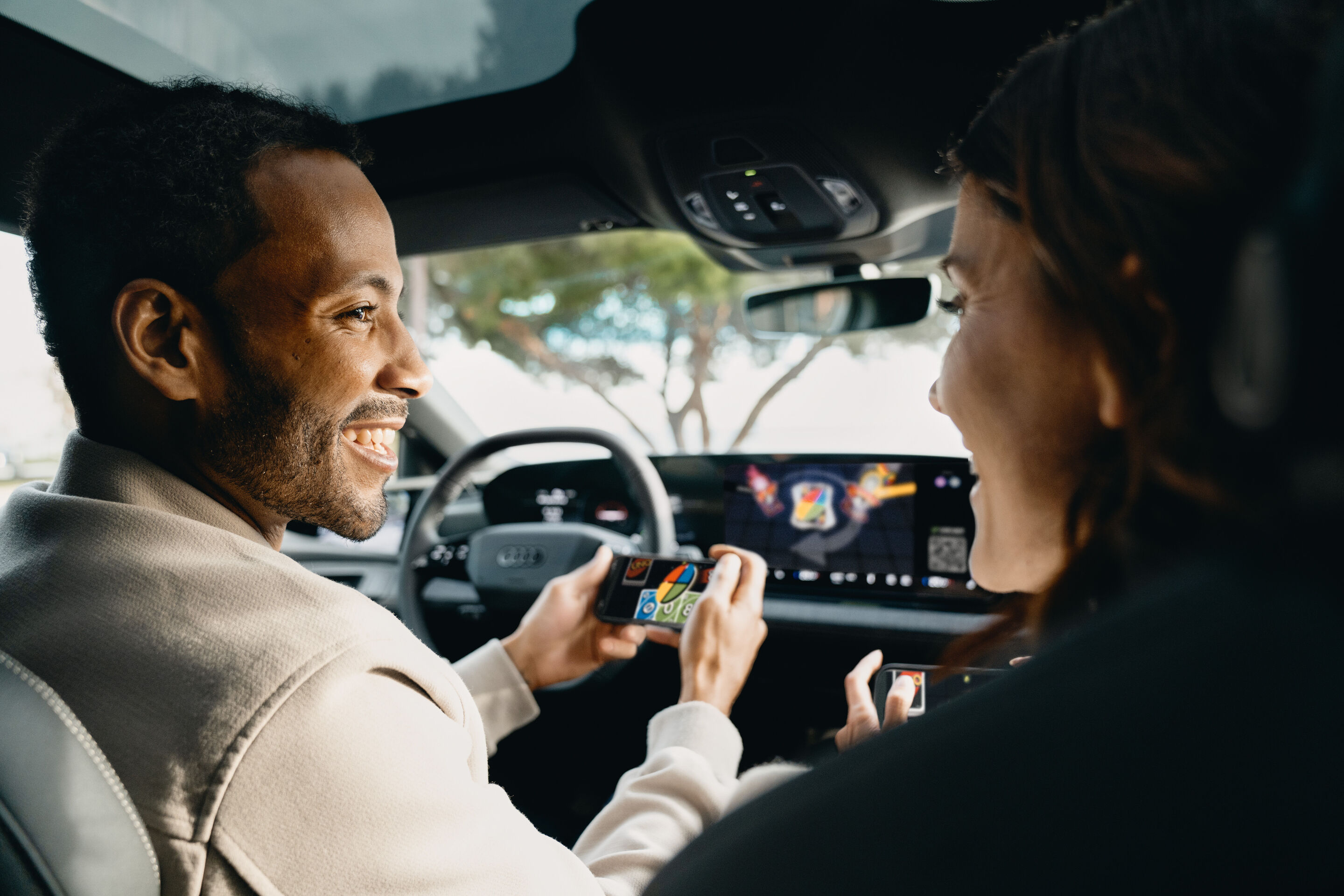 Spielspaß unterwegs: Audi integriert AirConsole - Auf dem Touchdisplay in einem Audi läuft ein Spiel der Gaming-Plattform AirConsole. Fahrer und Beifahrerin lächeln einander an und halten je ein Smartphone in ihrer Hand, welches sie als Controller zum...