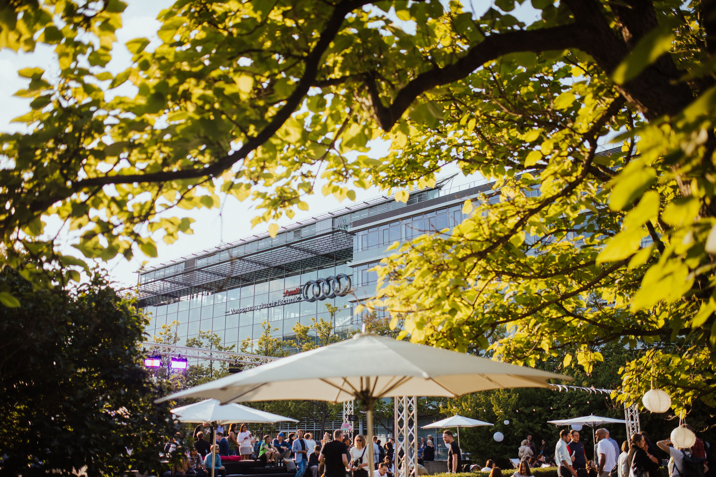 Audi Summer Sounds: Musik, Drinks und Streetfood am Audi Forum - Im Audi Forum Ingolstadt sind bei den Audi Summer Sounds 2024 im Außenbereich Sonnenschirme aufgespannt, unter denen sich zahlreiche Menschen aufhalten.
