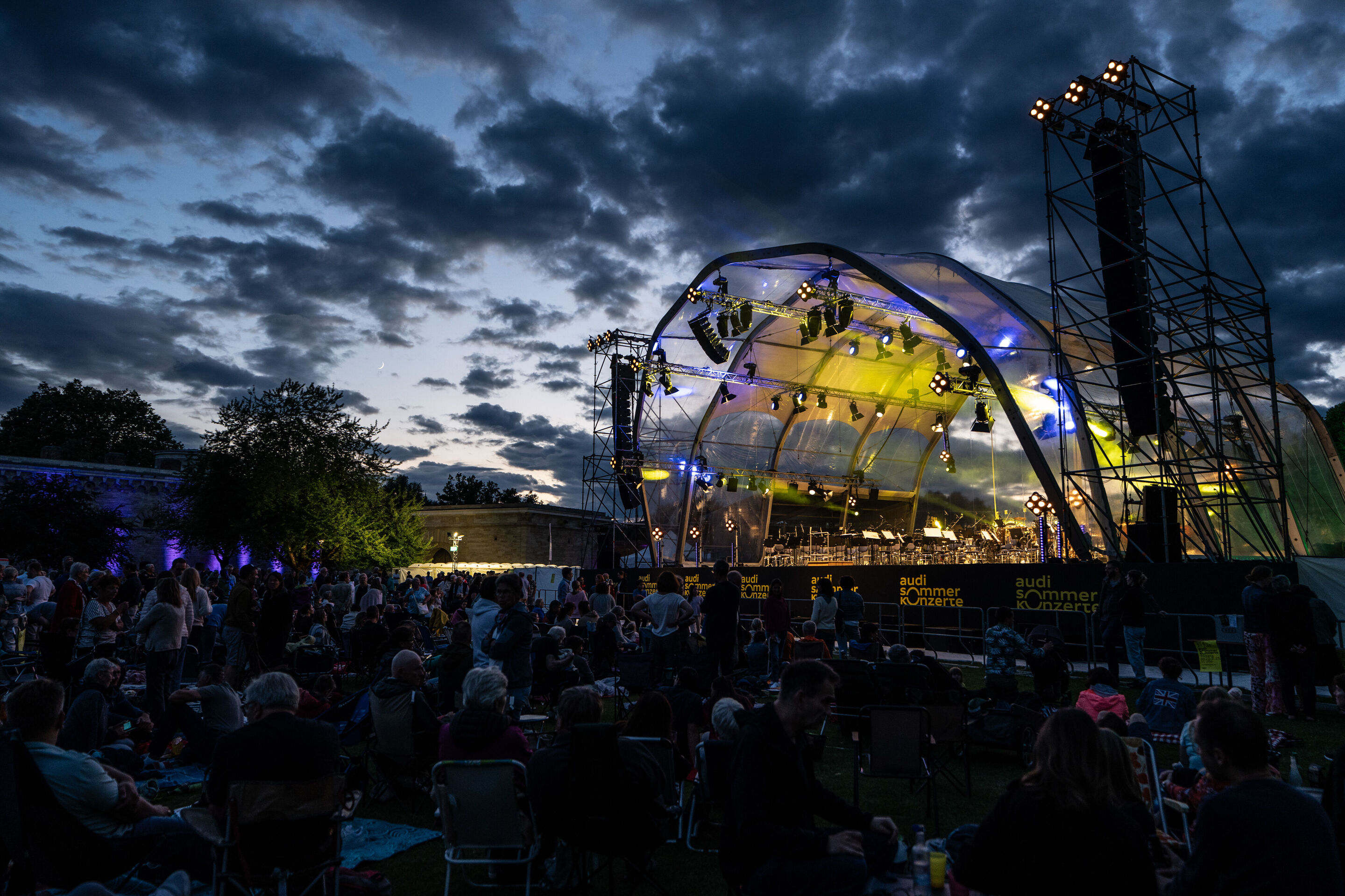 Classic Open Air – musical highlights with the Audi Philharmonic Wind Orchestra - At the Audi Summer Concerts Classic Open Air event in Klenzepark, the audience sits in front of the illuminated stage at dusk.