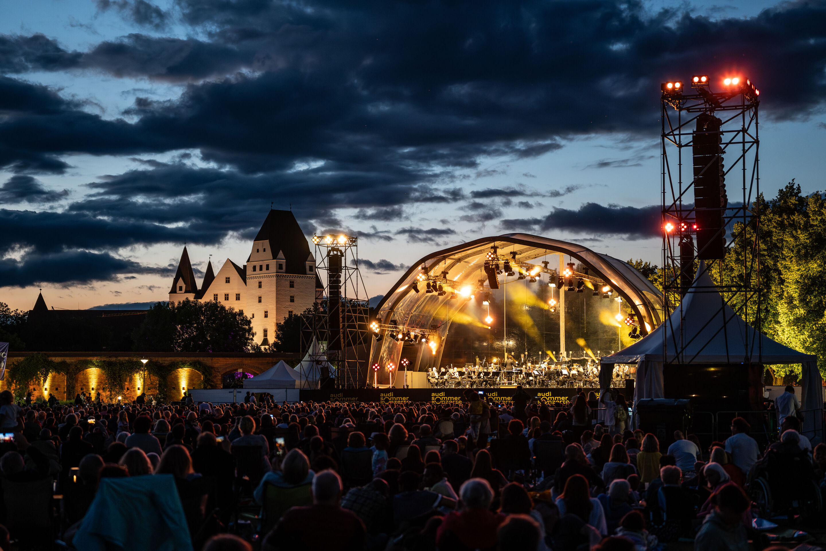 Classic Open Air – musical highlights with the Audi Philharmonic Wind Orchestra - At the Audi Summer Concerts Classic Open Air event in Klenzepark, the audience sits in front of the illuminated stage at dusk.