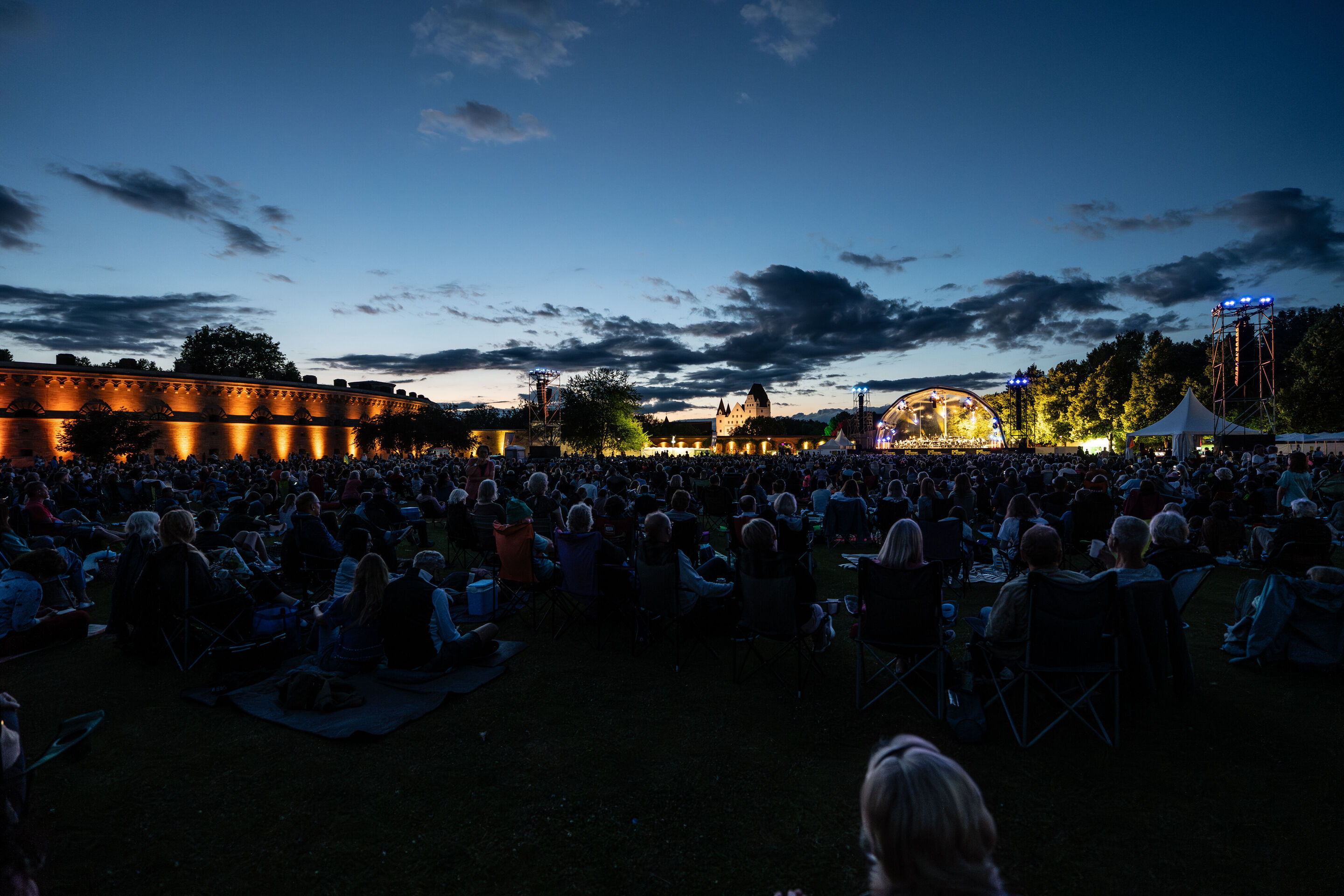 Classic Open Air – musical highlights with the Audi Philharmonic Wind Orchestra - At the Audi Summer Concerts Classic Open Air event in Klenzepark, the audience sits in front of the illuminated stage at dusk; on the left side, the historical structure...