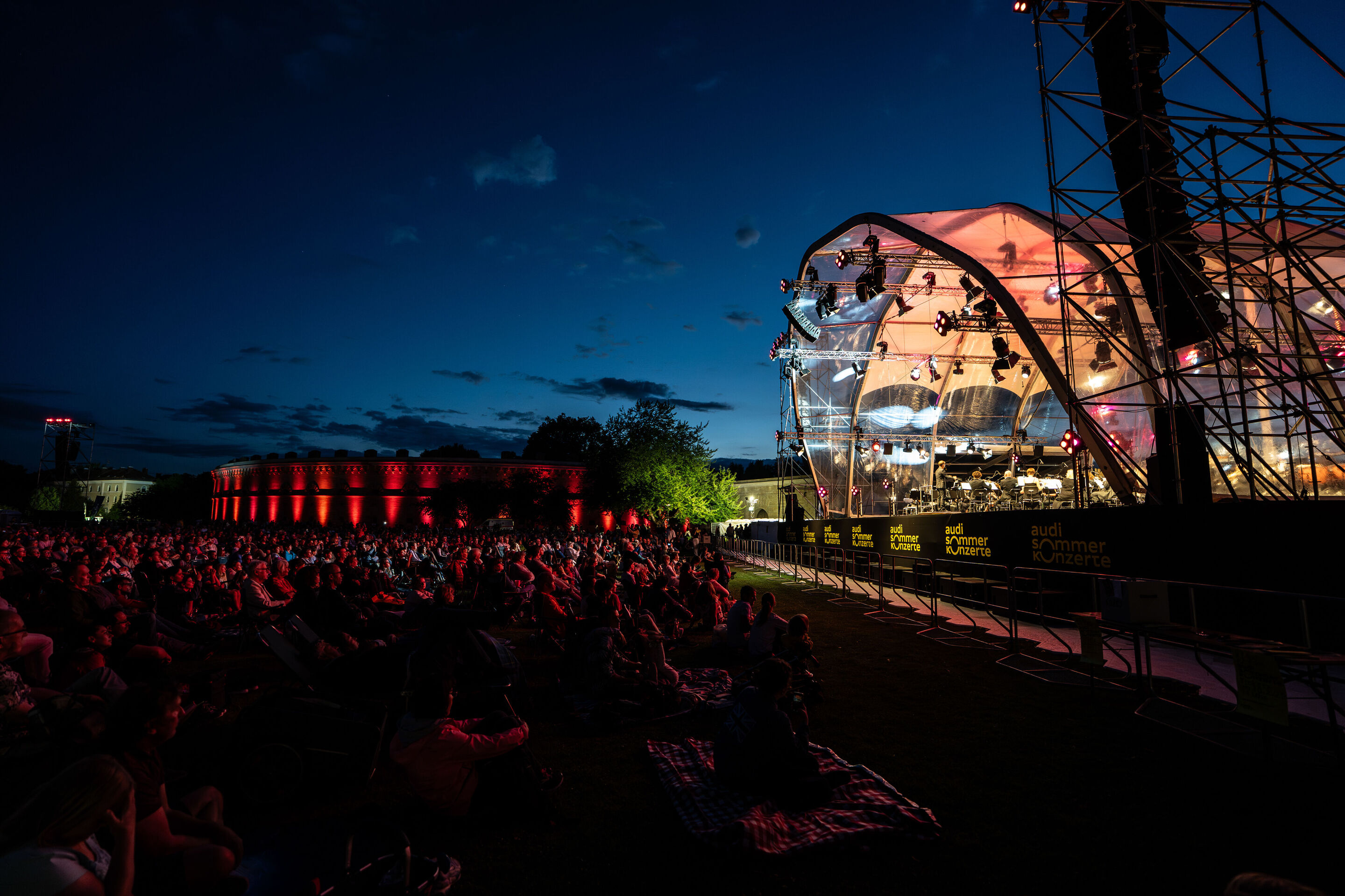 Classic Open Air – musical highlights with the Audi Philharmonic Wind Orchestra - At the Audi Summer Concerts Classic Open Air event in Klenzepark, the audience sits in front of the illuminated stage at dusk; on the left side, the historical structure...