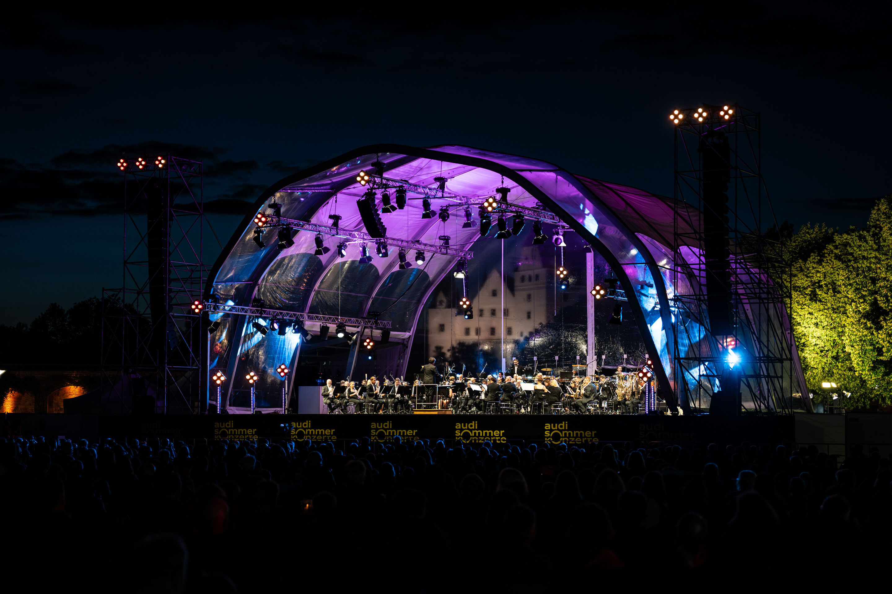 Classic Open Air – musical highlights with the Audi Philharmonic Wind Orchestra - At the Audi Summer Concerts Classic Open Air event in Klenzepark, the audience sits in front of the illuminated stage in the evening.