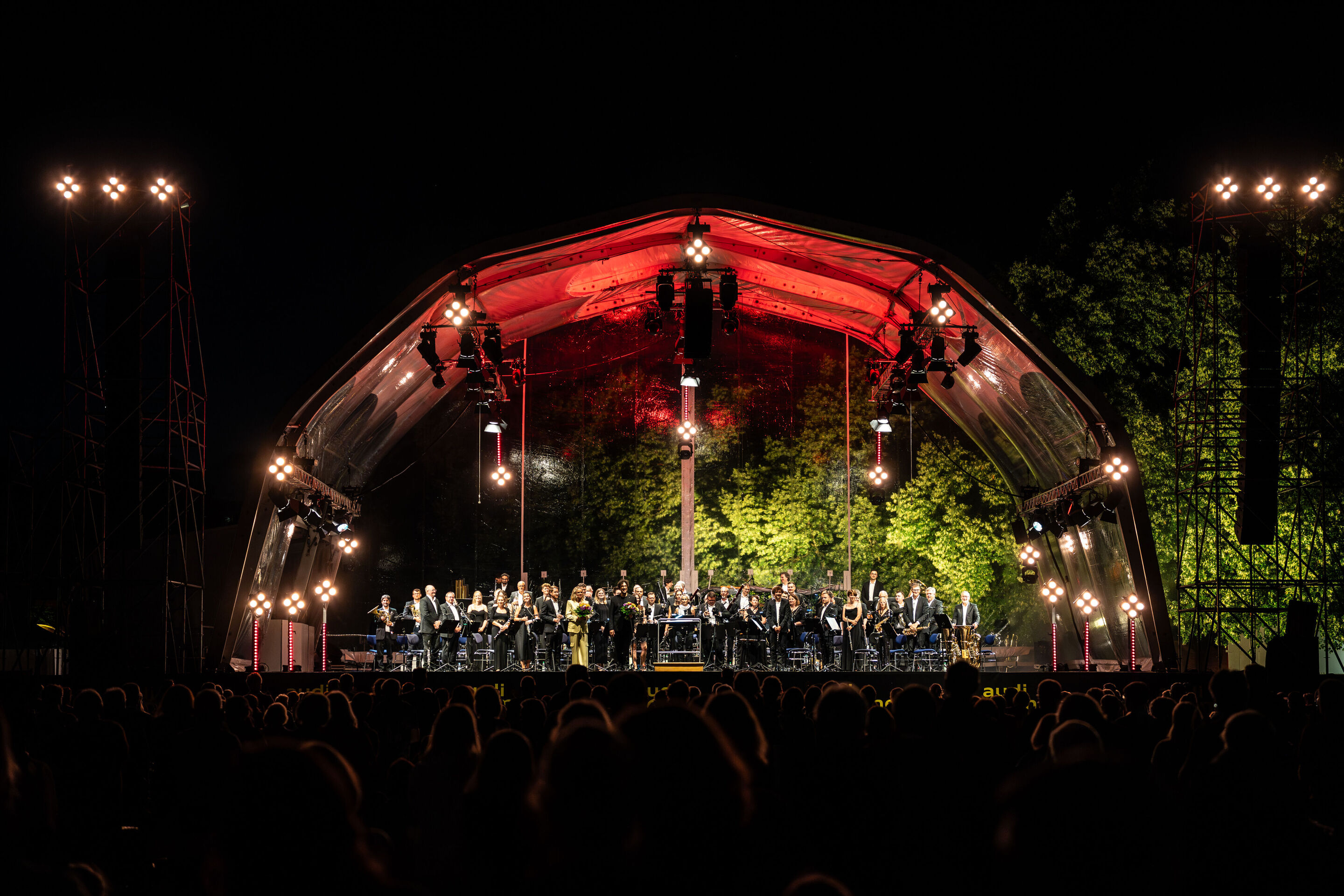 Classic Open Air – musical highlights with the Audi Philharmonic Wind Orchestra - The Audi Philharmonic Wind Orchestra, conductor Pietro Sarno, and host Annekatrin Hentschel stand on the illuminated stage at the Classic Open Air event in Klenzepark as...