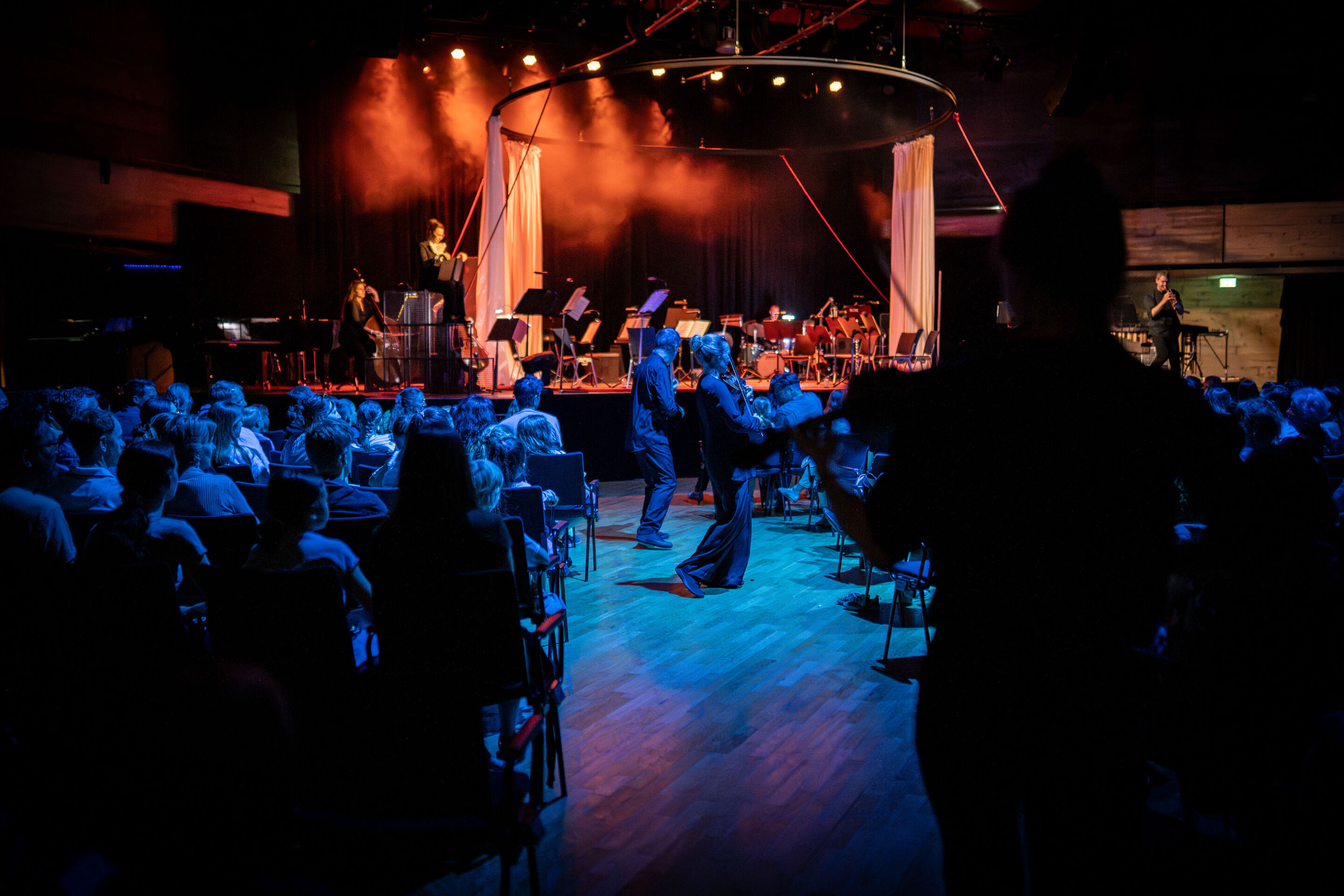 „Carnival“: Inszeniertes Familienkonzert rund um die fabelhafte Welt der Instrumente - Im Festsaal Ingolstadt musizieren einige Mitglieder des Aurora Orchestra zwischen dem Publikum im Stehen. Im Hintergrund auf der Bühne ist links Erzählerin Birgit Minichmayr...
