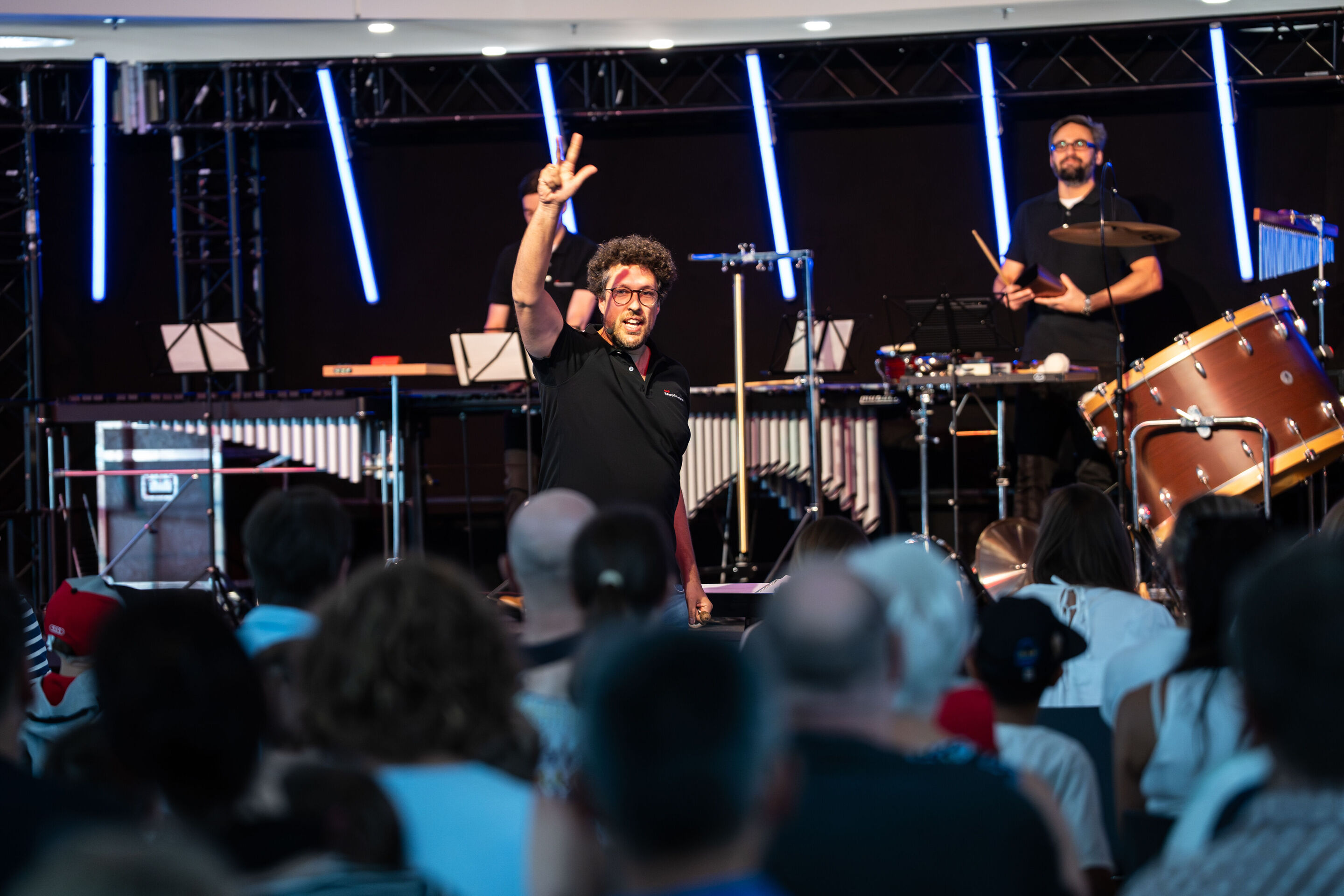 „ADUI und die Kraft der magischen Ringe“: Kinderkonzert mit der Audi Bläserphilharmonie - Dirigent Pietro Sarno auf der Bühne im Gebäude H6 am Standort Ingolstadt stehend, blickt auf das Publikum und hält drei Finger hoch.