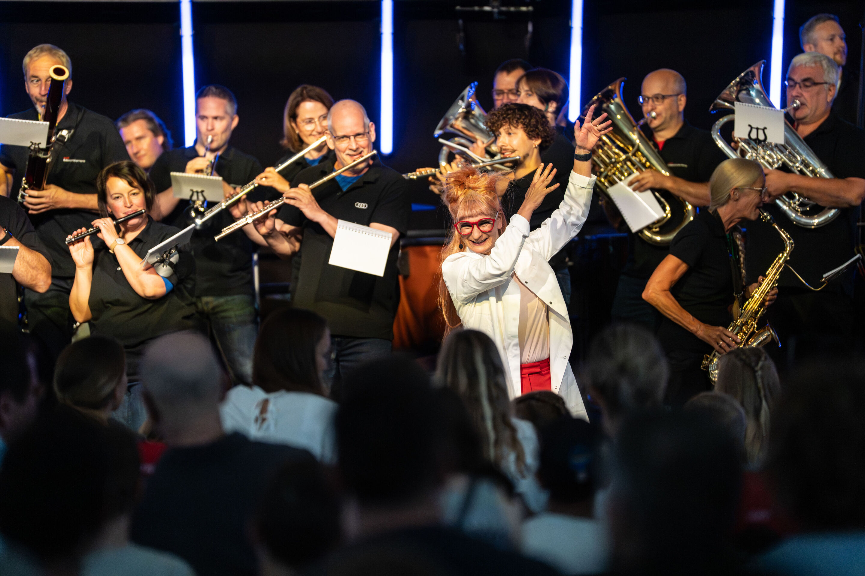 „ADUI und die Kraft der magischen Ringe“: Kinderkonzert mit der Audi Bläserphilharmonie - Aus Sicht des Publikums ist im H6  Forscherin Olivia Wendt in der Mitte zu sehen, hinter ihr die Musizierenden der Audi Bläserphilharmonie.