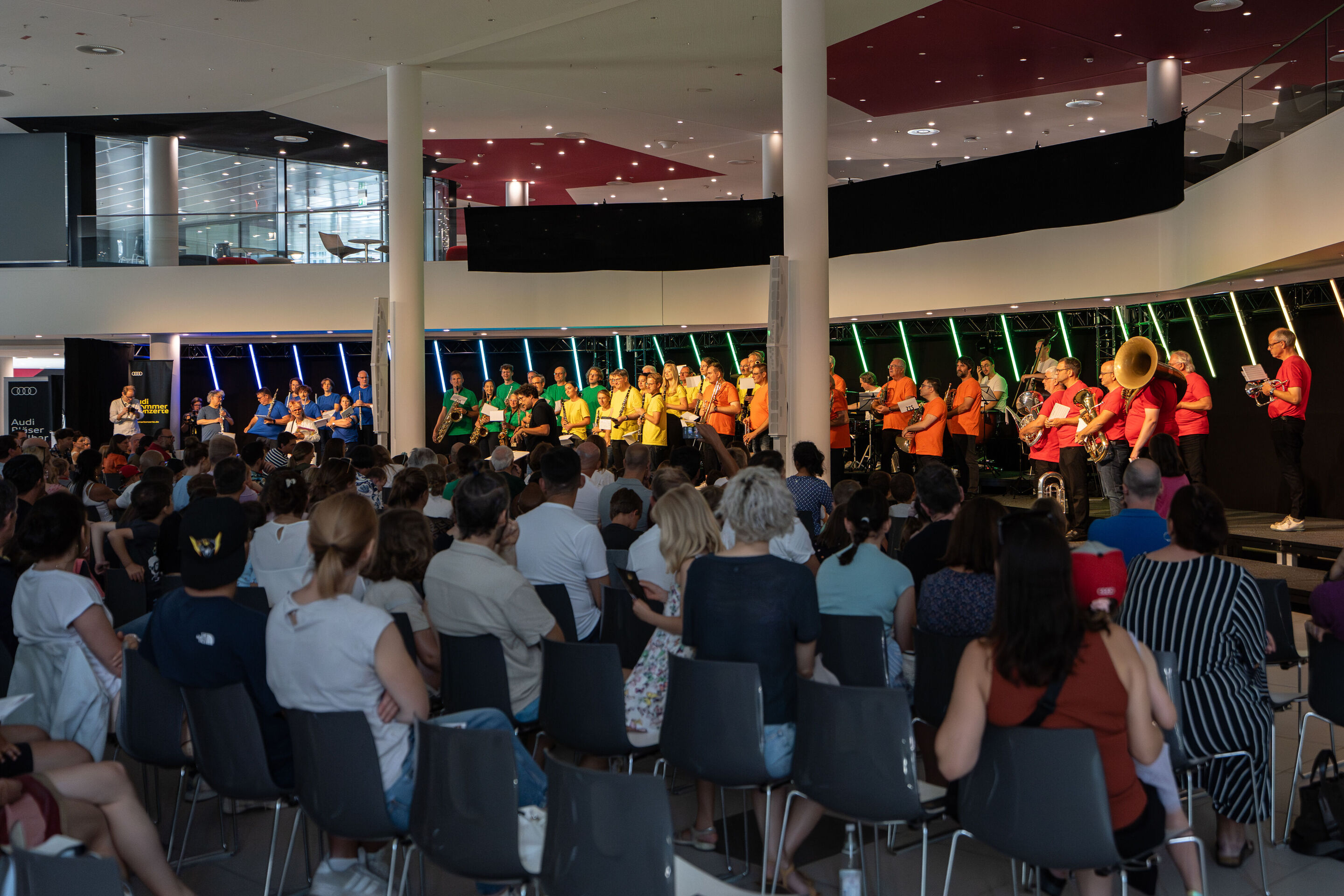 „ADUI und die Kraft der magischen Ringe“: Kinderkonzert mit der Audi Bläserphilharmonie - Die Musizierende der Audi Bläserphilharmonie sind in bunten T-Shirts gekleidet mit Dirigent Pietro Sarno in der Mitte auf der Bühne des H6 Gebäudes am Standort Ingolstadt.