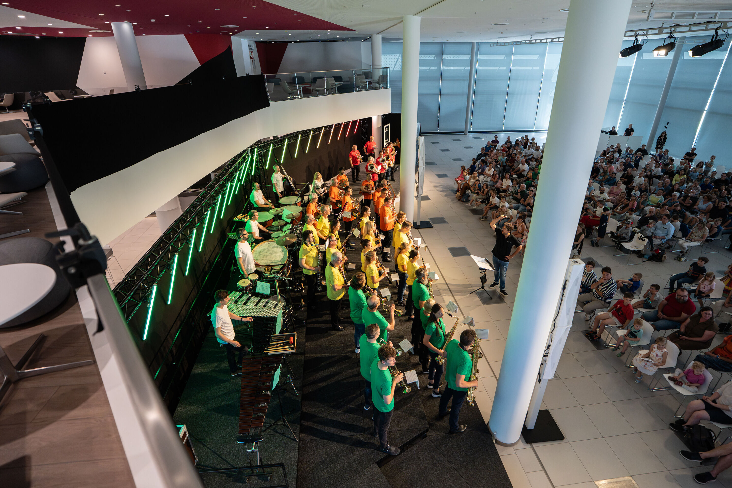 „ADUI und die Kraft der magischen Ringe“: Kinderkonzert mit der Audi Bläserphilharmonie - Auf der Bühne im H6 sind von oben die Musiziereden der Audi Bläserphilharmonie mit Dirigent Pietro Sarno und das Publikum zu sehen.