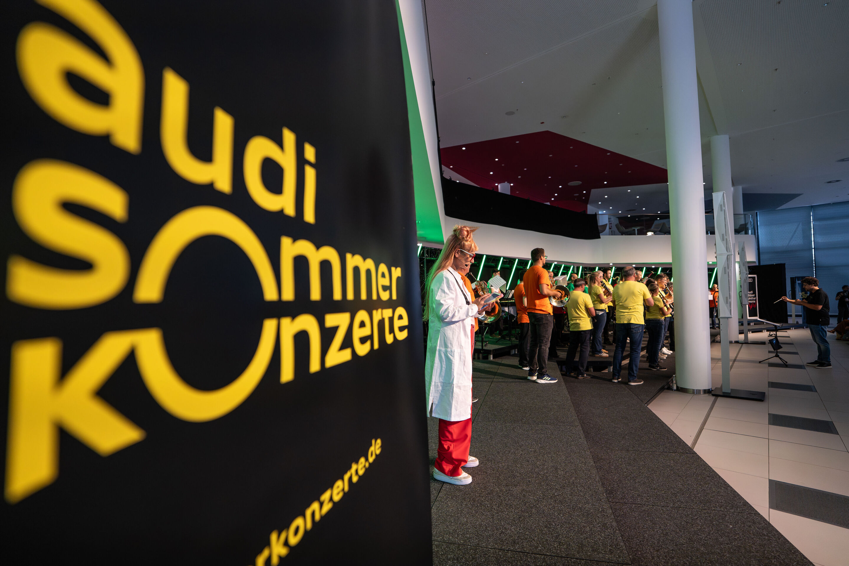 „ADUI und die Kraft der magischen Ringe“: Kinderkonzert mit der Audi Bläserphilharmonie - Im Vordergrund steht Olivia Wendt im Forscherkostüm, während das Orchester im Hintergrund spielt. Links außen ist das Logo der Audi Sommerkonzerte angebracht.