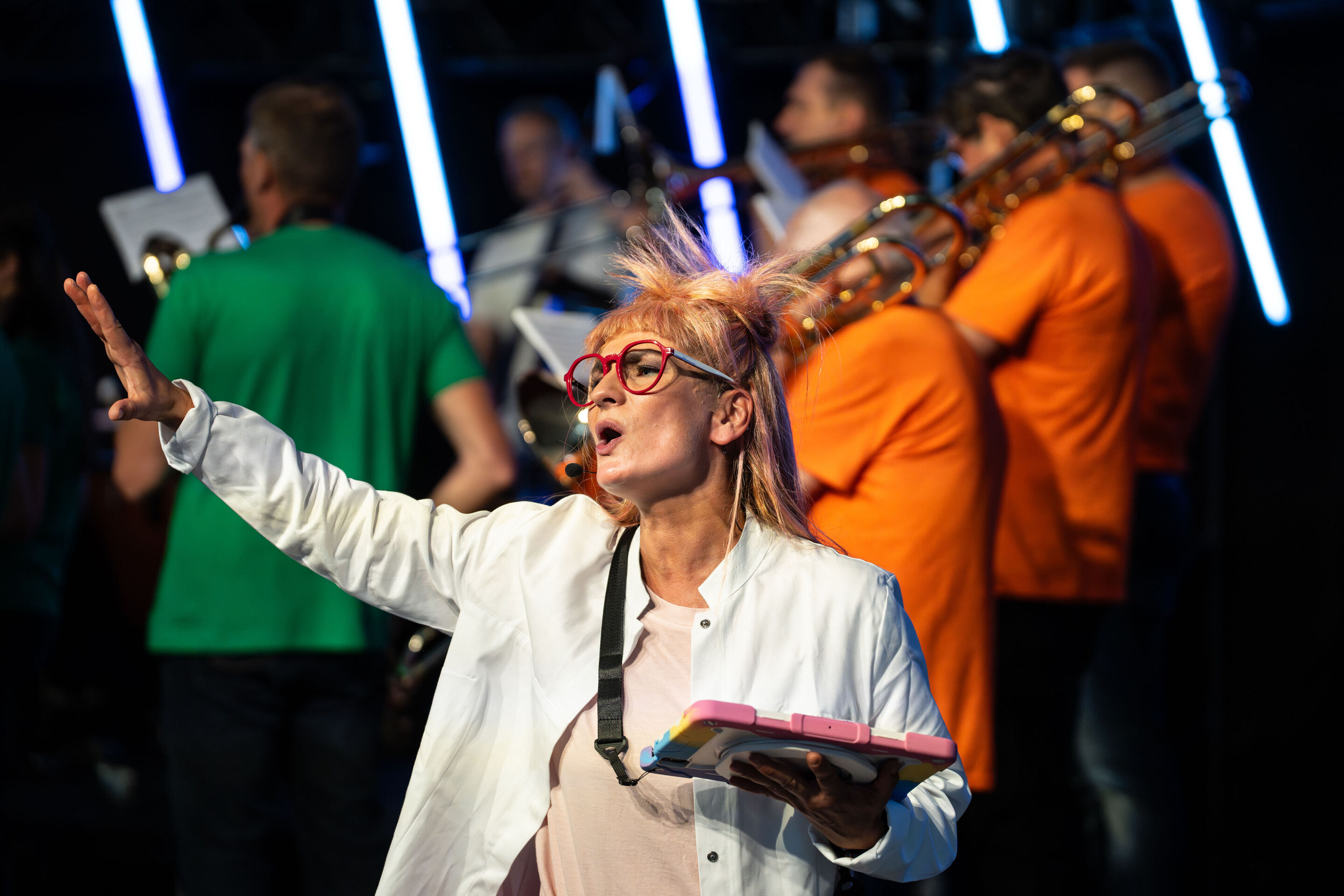 „ADUI und die Kraft der magischen Ringe“: Kinderkonzert mit der Audi Bläserphilharmonie - Olivia Wendt steht im weißen Kittel als Forscherin auf der Bühne, hält ein Tablet in der Hand und gestikuliert. Im Hintergrund spielt die Audi Bläserphilharmonie,...
