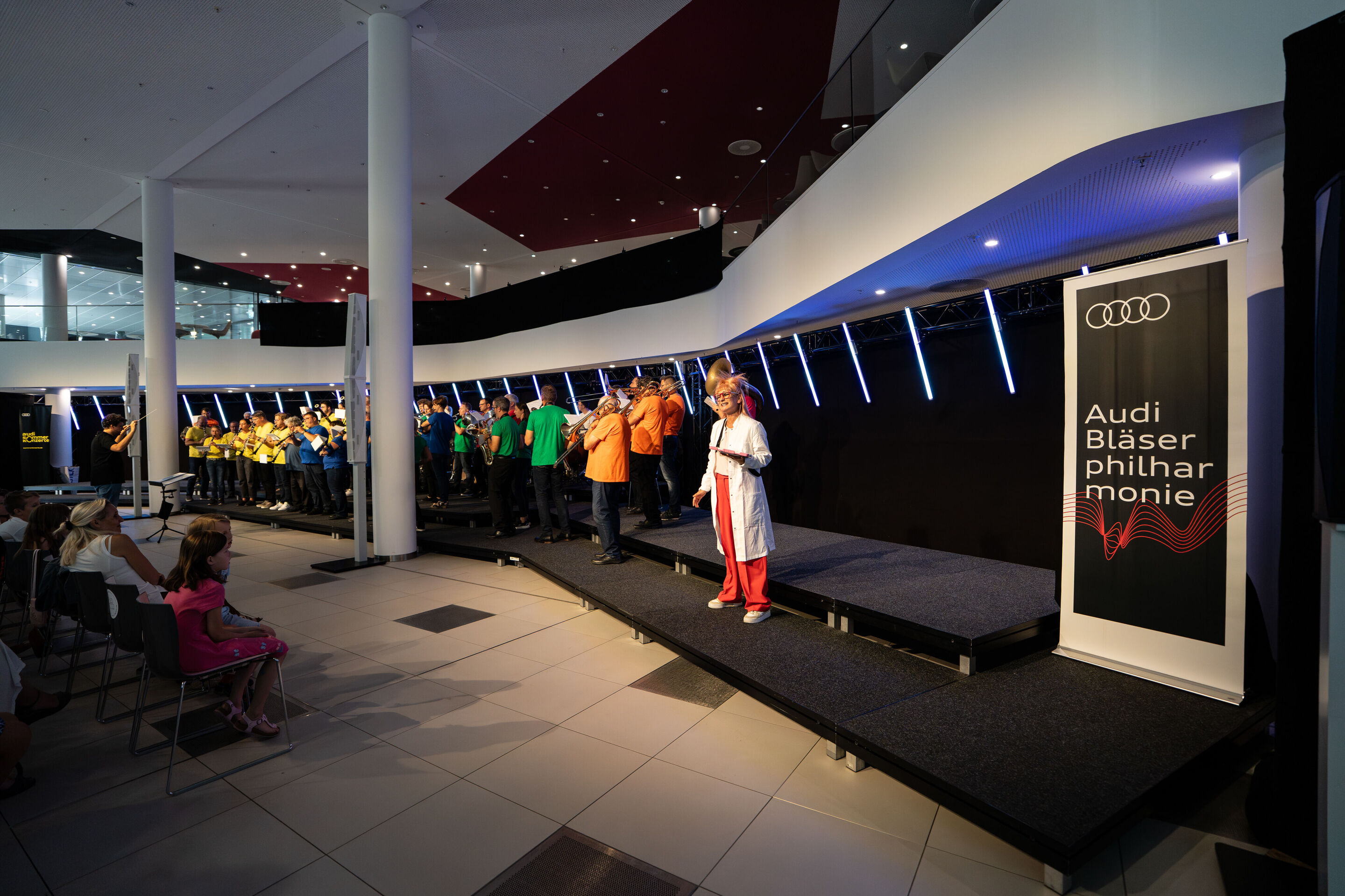 „ADUI und die Kraft der magischen Ringe“: Kinderkonzert mit der Audi Bläserphilharmonie - Auf der Bühne steht Olivia Wendt in einem Wissenschaftler-Kostüm. Im Hintergrund spielt die Audi Bläserphilharmonie unter der Leitung von Dirigent Pietro Sarno.
