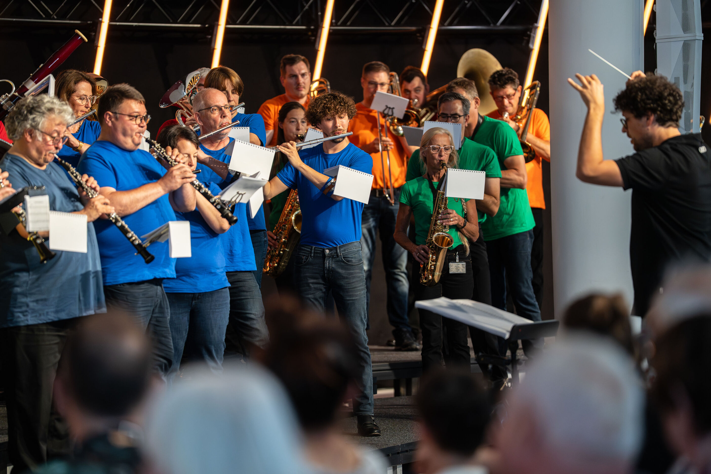 „ADUI und die Kraft der magischen Ringe“: Kinderkonzert mit der Audi Bläserphilharmonie - Die Audi Bläserphilharmonie spielt auf der Bühne. Rechts im Bild ist Dirigent Pietro Sarno zu sehen. Die Musiker tragen bunte T-Shirts und spielen verschiedene Blasinstrumente.