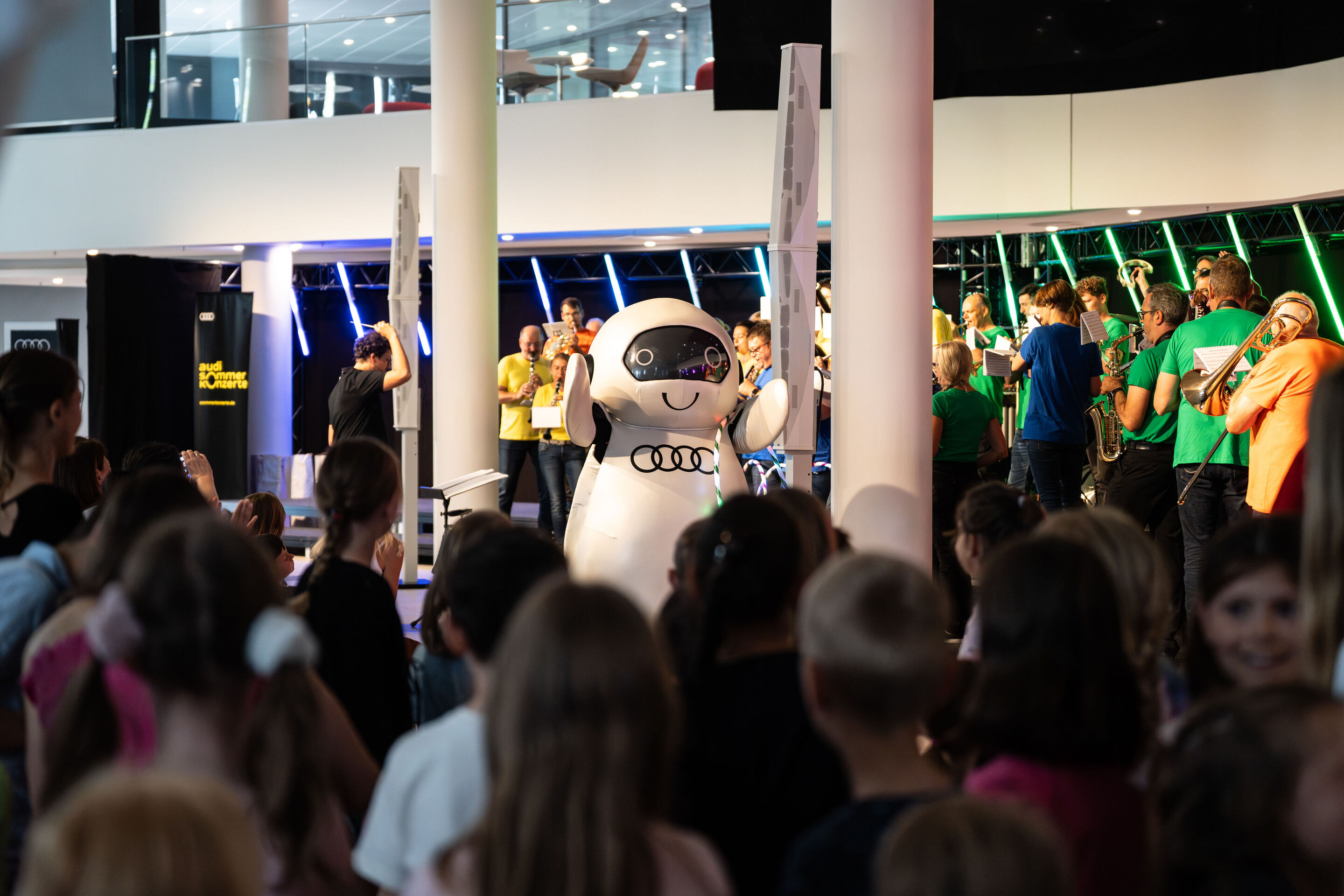 „ADUI und die Kraft der magischen Ringe“: Kinderkonzert mit der Audi Bläserphilharmonie - Der Roboter ADUI mit den vier Audi-Ringen steht vor der Bühne. Im Hintergrund spielt die Audi Bläserphilharmonie unter der Leitung von Pietro Sarno vor dem Publikum.