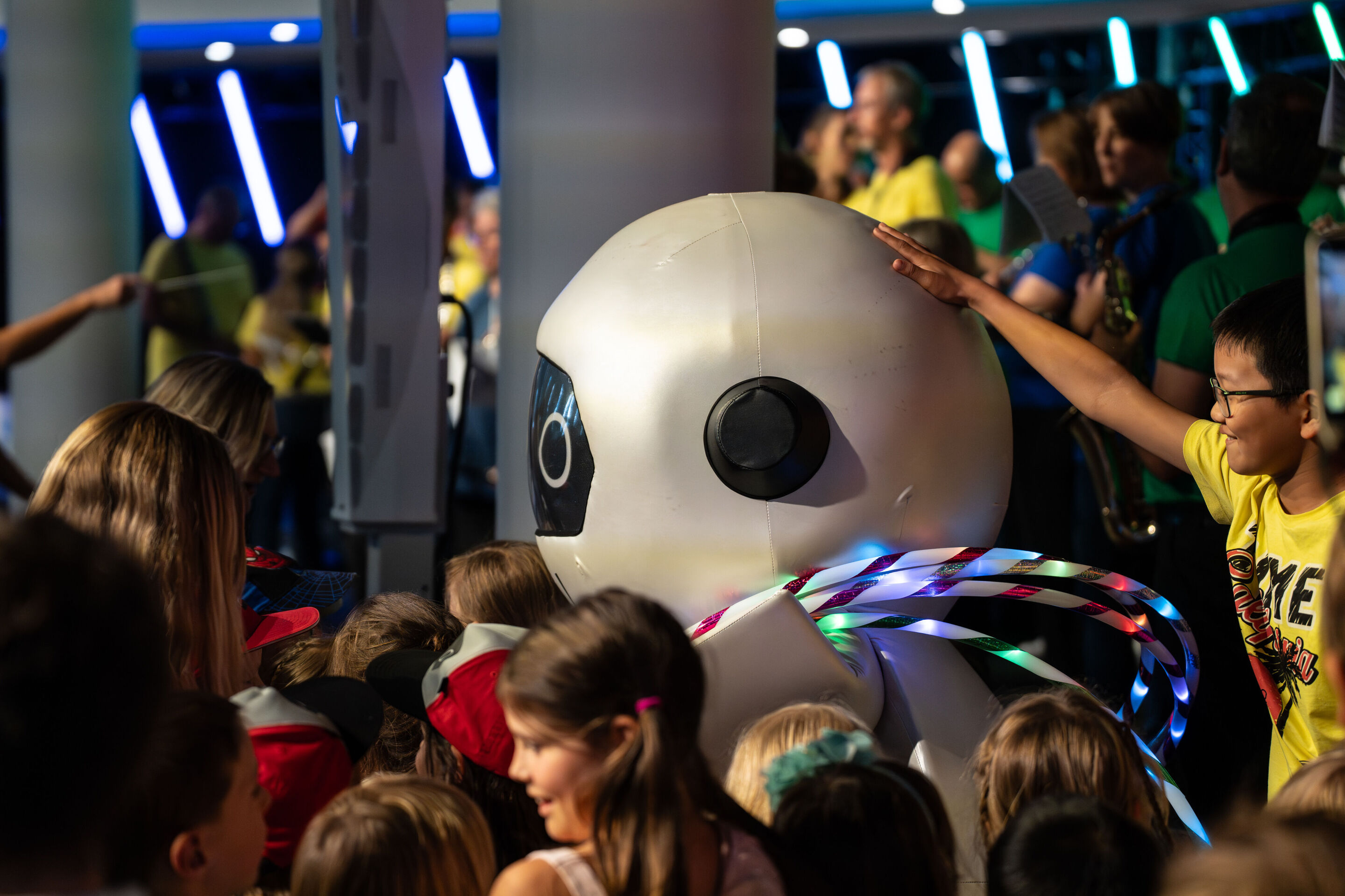 „ADUI und die Kraft der magischen Ringe“: Kinderkonzert mit der Audi Bläserphilharmonie - Viele Kinder stehen um den Roboter ADUI. Im Hintergrund spielt die Audi Bläserphilharmonie.
