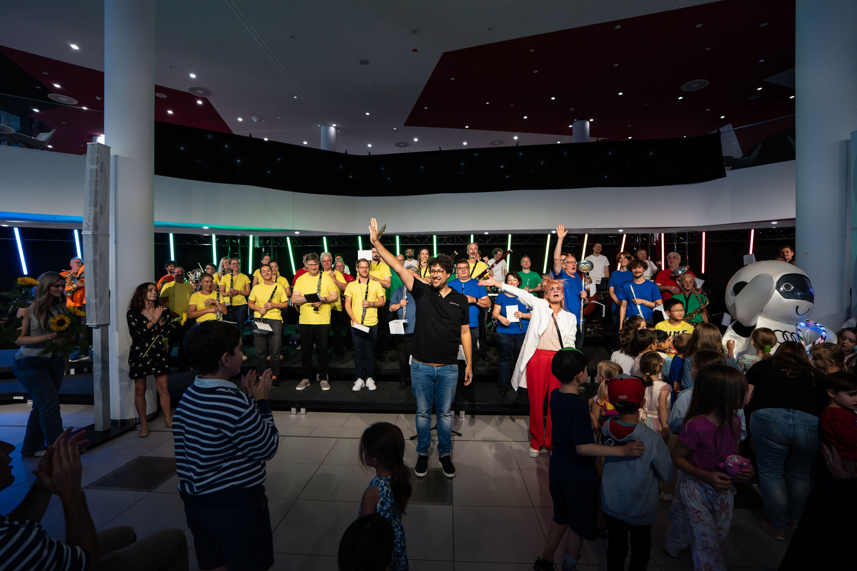 „ADUI und die Kraft der magischen Ringe“: Kinderkonzert mit der Audi Bläserphilharmonie - Auf einer Bühne steht die Audi Bläserphilharmonie mit Dirigent Pietro Sarno in der Mitte und der Schauspielerin Olivia Wendt rechts von ihm vor dem Publikum und...