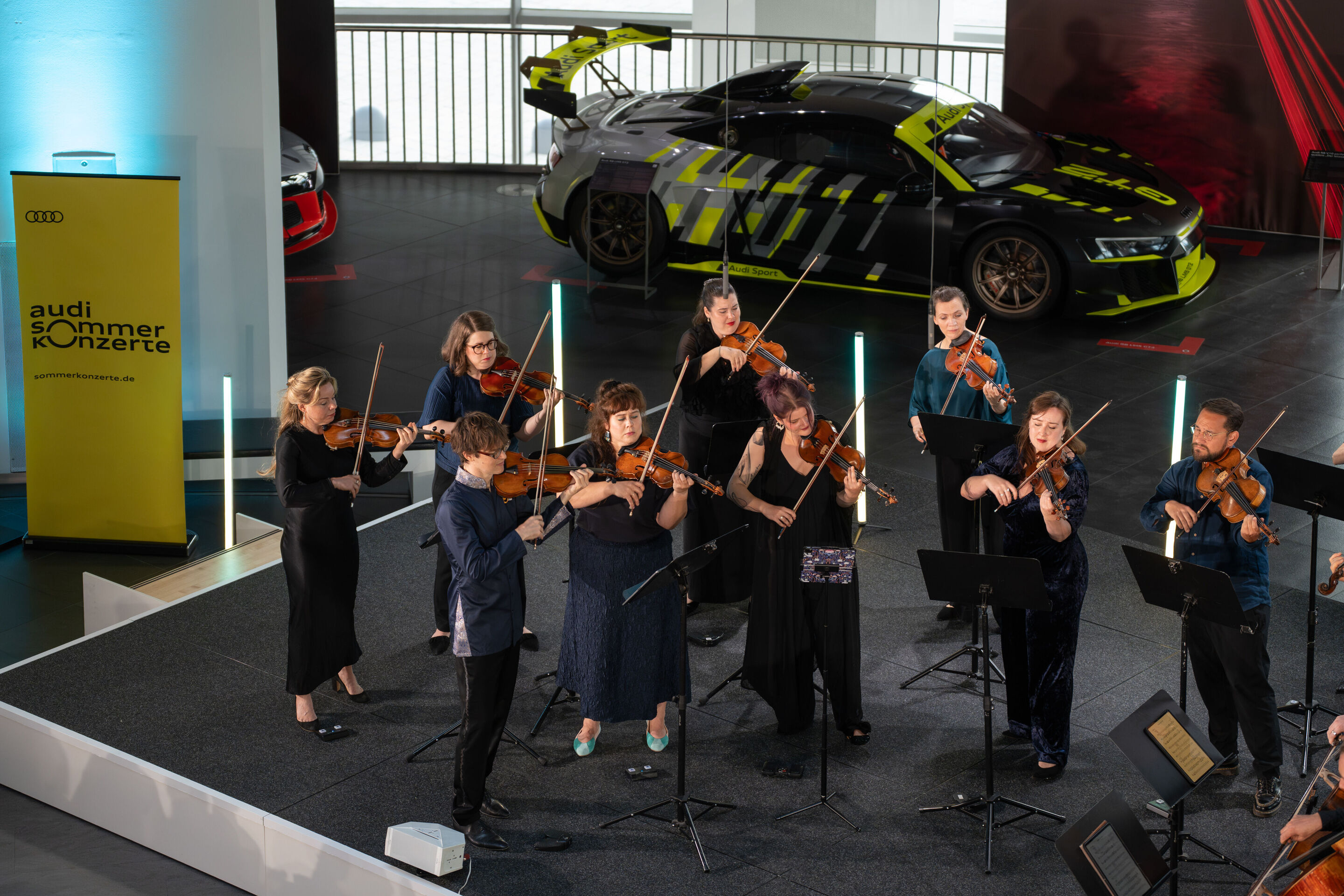 „Crossing over the Atlantic“: von Britten bis Nirvana mit dem O/Modernt Kammerorchester - Auf der Bühne des Audi museum mobile stehen die Geiger des O Modernt Kammerorchesters. Im Hintergrund ein Audi R8 als Ausstellungsstück der „Audi R8 - Generations...
