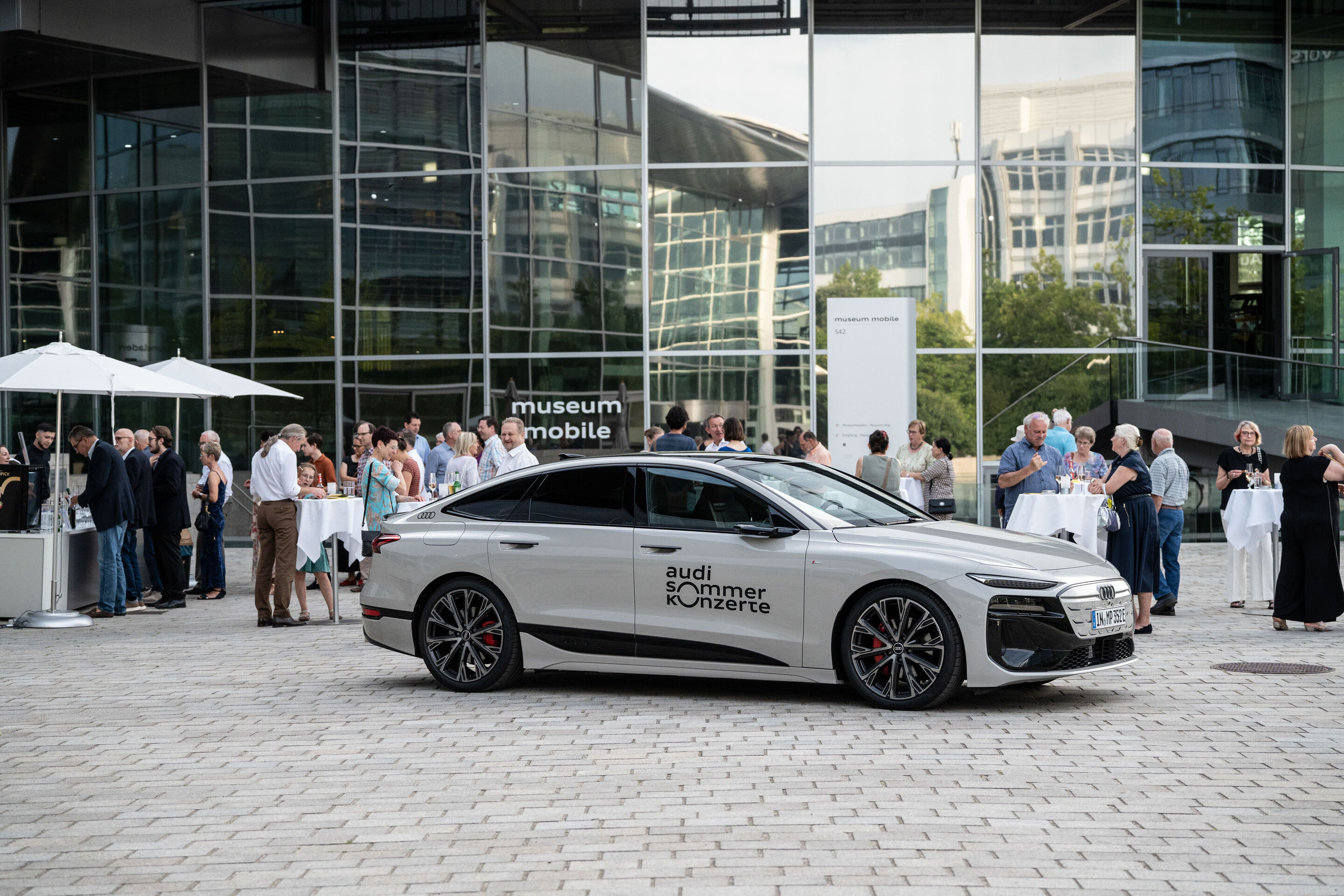 Alinde Quintet – winners of the ARD Music Competition - Ein Audi A6 Sportback e-tron in der Farbe Siambeige Metallic mit dem Logo der Audi Sommerkonzerte auf der Piazza vor dem museum mobile. Im Hintergrund: Gäste der Audi Sommerkonzerte.