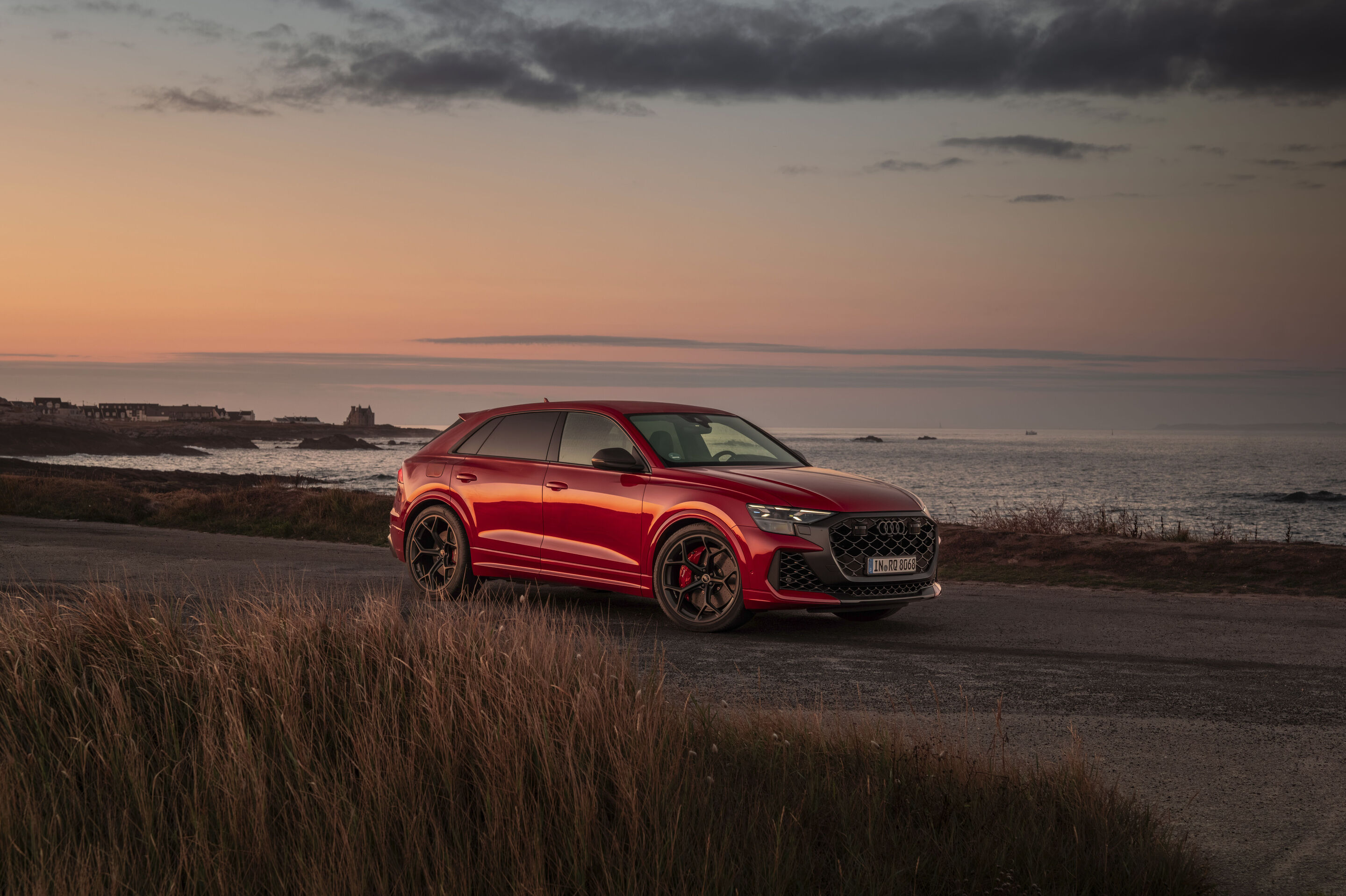 Audi RS Q8 SUV performance - Audi RS Q8 SUV performance in Chilirot Metallic auf einer Küstenstraße vor der Kulisse eines Sonnenuntergangs über dem Meer.
