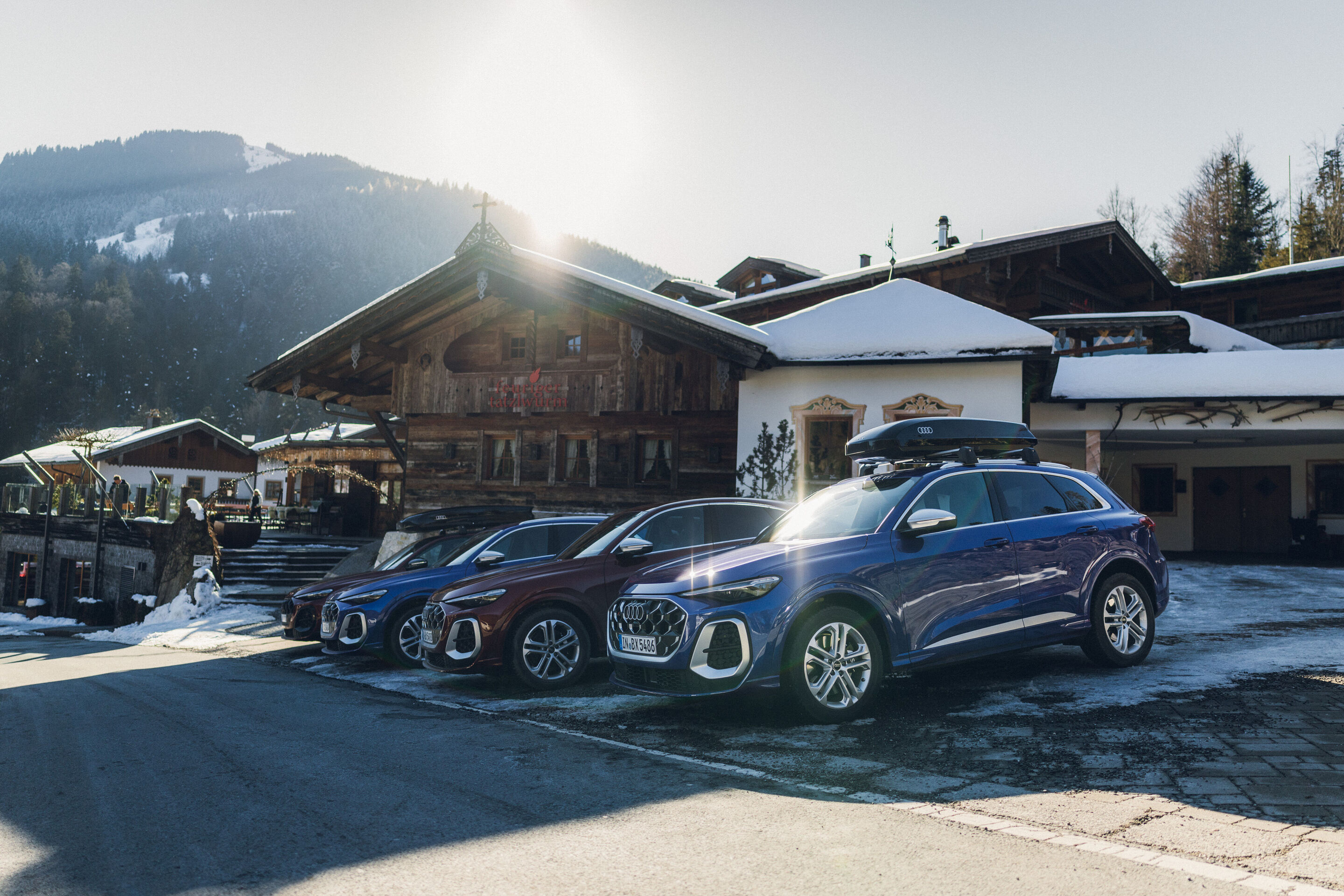 Audi SQ5 und Audi SQ5 Sportback - Vier Audi SQ5 Modelle stehen nebeneinander vor einem traditionellen alpinen Holzgebäude. Die Fahrzeuge in Ultrablau, Grenadinerot und Daytonagrau sind auf einem leicht verschneiten Hof geparkt; das vorderste und das hinterste...