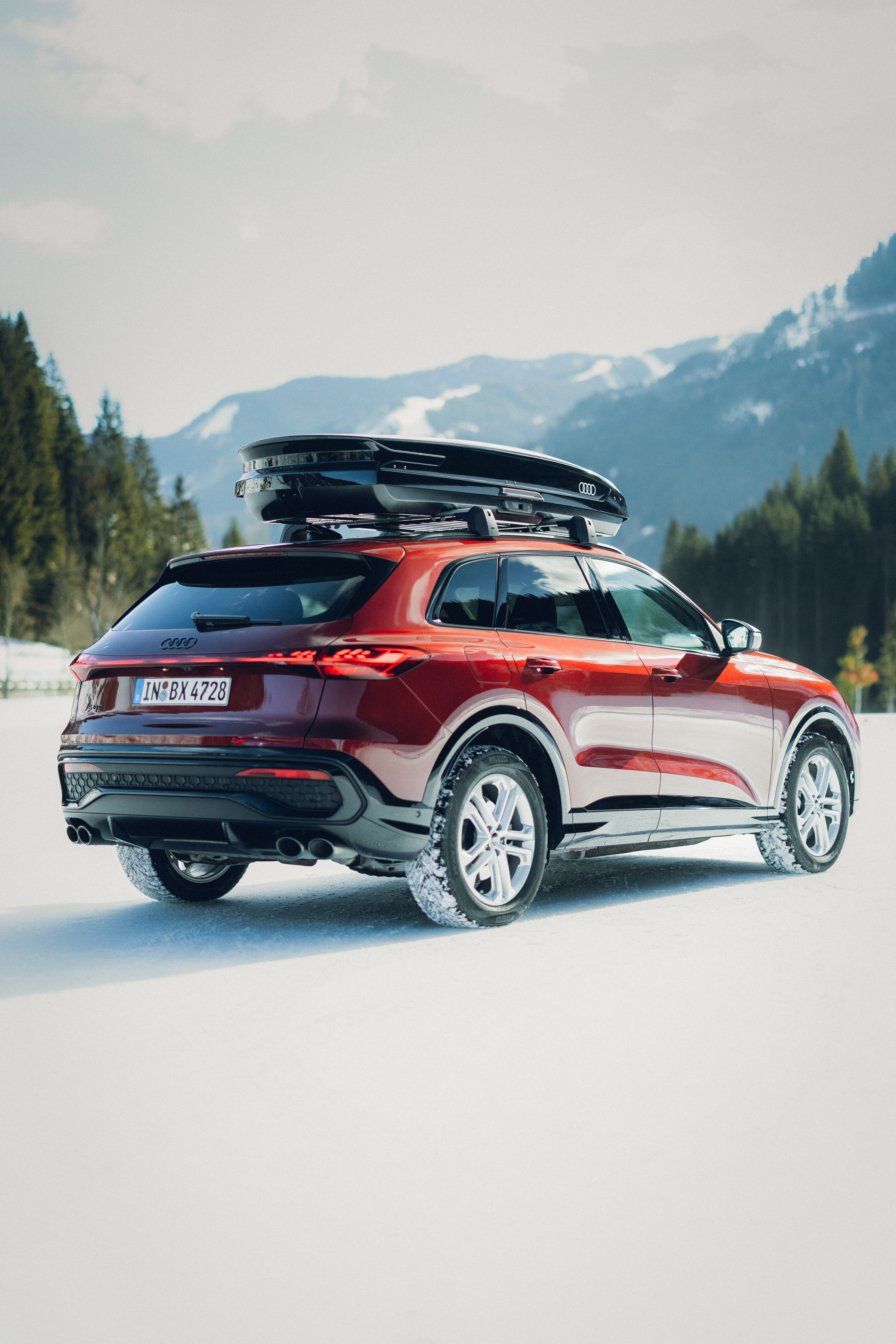 Audi SQ5 - Ein Audi SQ5 in Grenadinerot steht auf einer verschneiten Fläche vor einer Berglandschaft. Das Fahrzeug ist von hinten und seitlich zu sehen und trägt eine schwarze Dachbox.