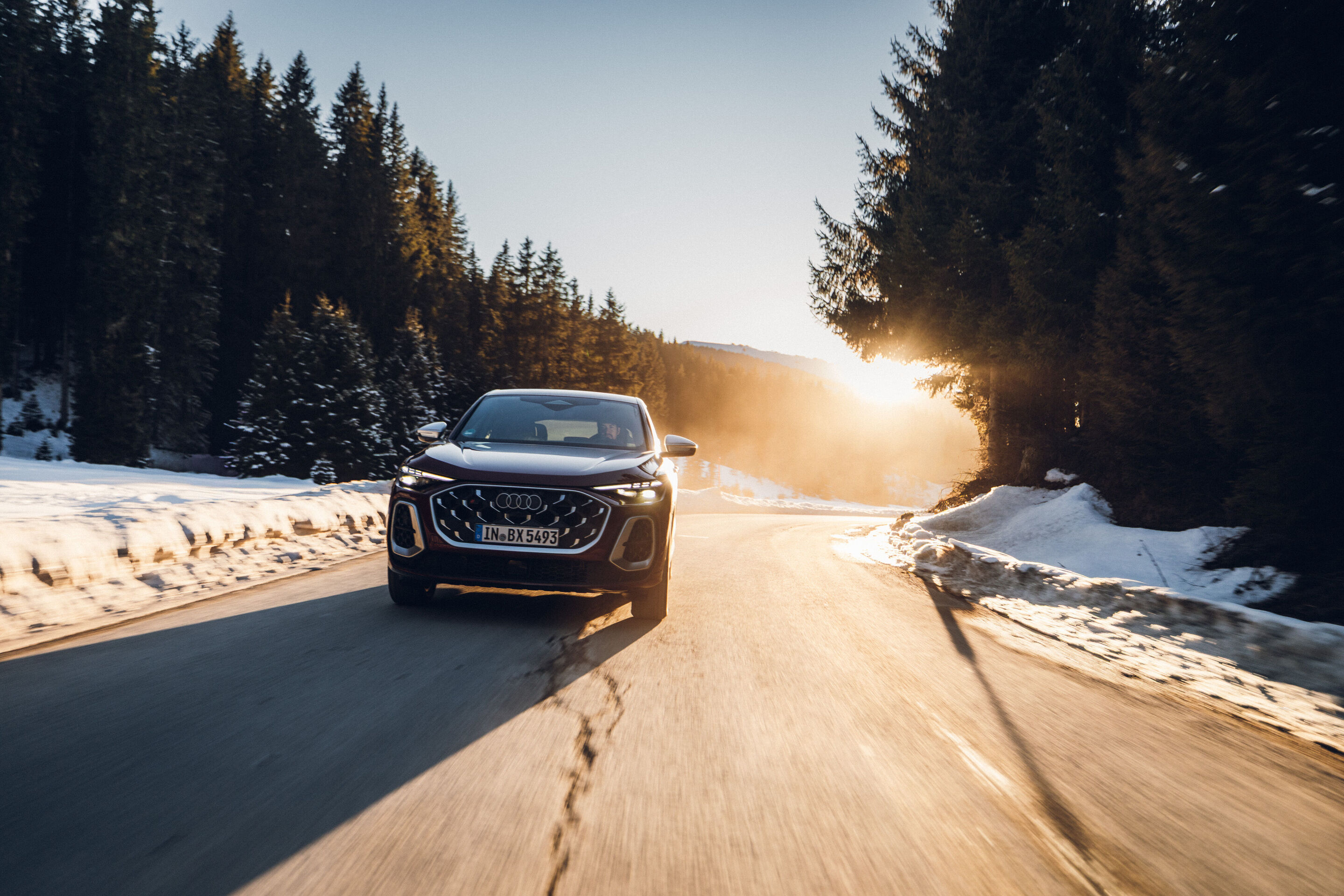 Audi SQ5 Sportback - Ein Audi SQ5 Sportback in Grenadinerot fährt auf einer winterlichen Bergstraße. Die Straße ist von Schnee gesäumt, und die Sonne scheint tief zwischen den Bäumen hindurch.