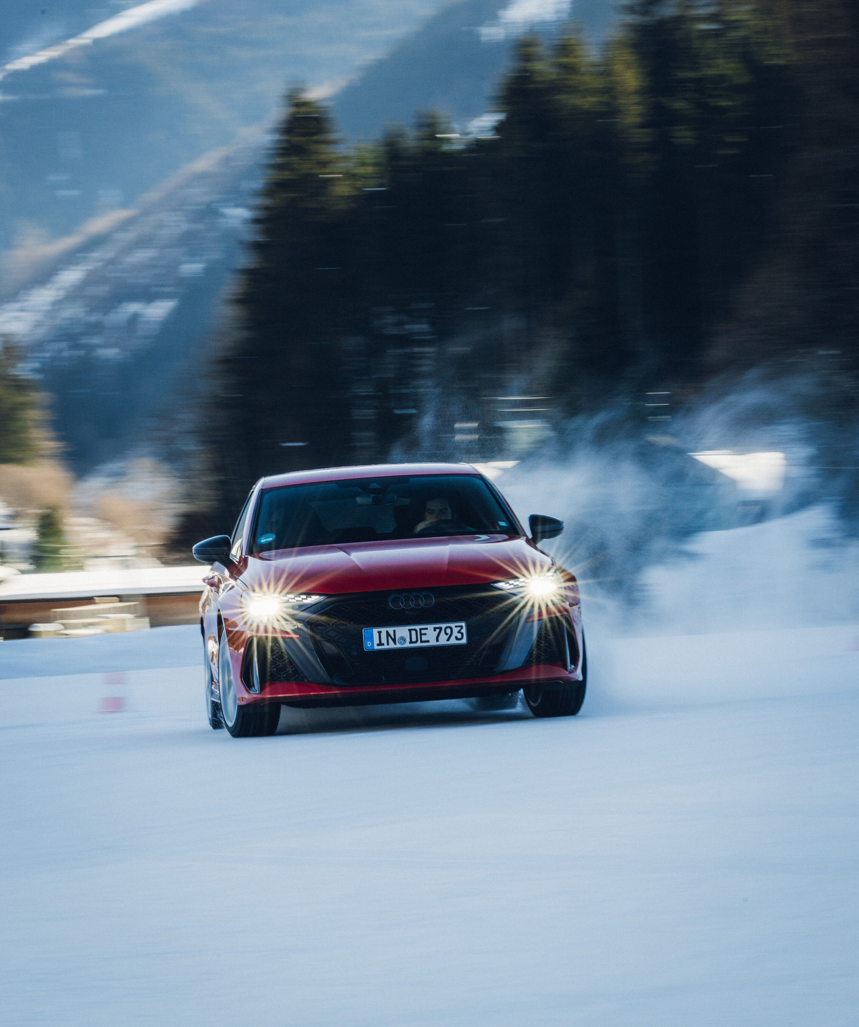 Audi RS 3 Sportback - Ein Audi RS 3 Sportback in Progressivrot fährt auf einer verschneiten Fläche und wirbelt Schnee auf. Das Auto ist frontal zu sehen, mit eingeschalteten Scheinwerfern.
