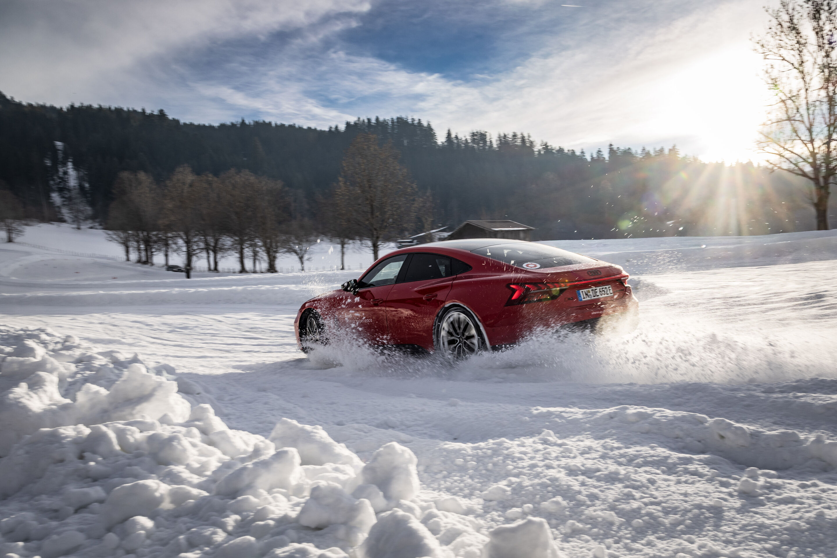 Audi RS e-tron GT - Der Audi RS e-tron GT fährt dynamisch über eine schneebedeckte Strecke und wirbelt dabei Schnee auf, im Hintergrund ein Wald unter der tief stehenden Sonne.
Farbe: Progressivrot Metallic, Outdoor Fahraufnahme, Exterieur, Dreiviertelheckansicht.