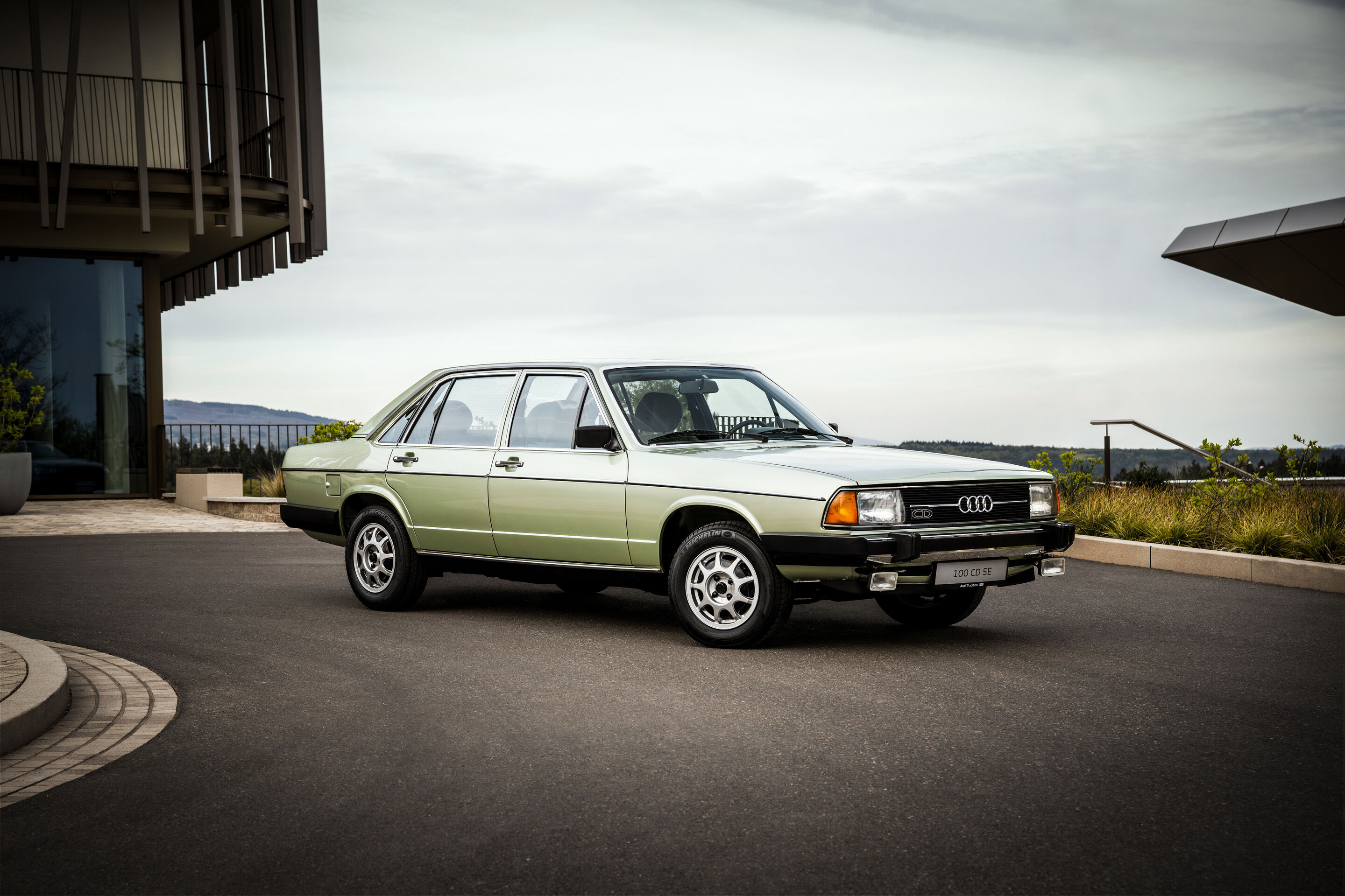 50 Jahre Fünfzylinder - Ein Audi 100 CD (C2, Baujahr 1979) in hellem Metallicgrün steht seitlich ausgerichtet auf einer befestigten Vorfläche vor moderner Architektur.