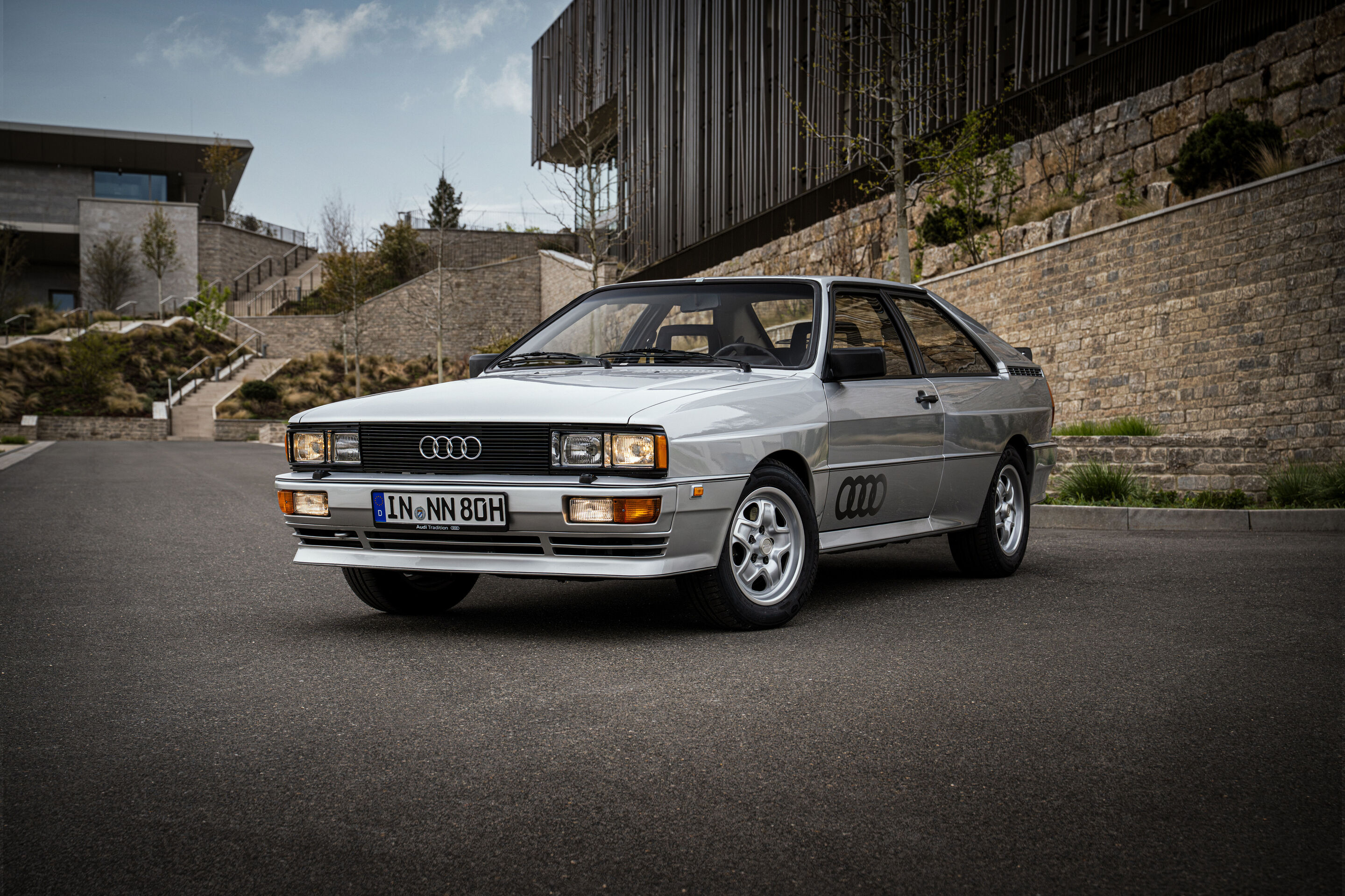 50 Jahre Fünfzylinder - Ein silberfarbener Audi quattro von 1980 mit verbreiterten Radhäusern ist in Dreiviertel-Frontansicht auf einer Asphaltfläche positioniert.
