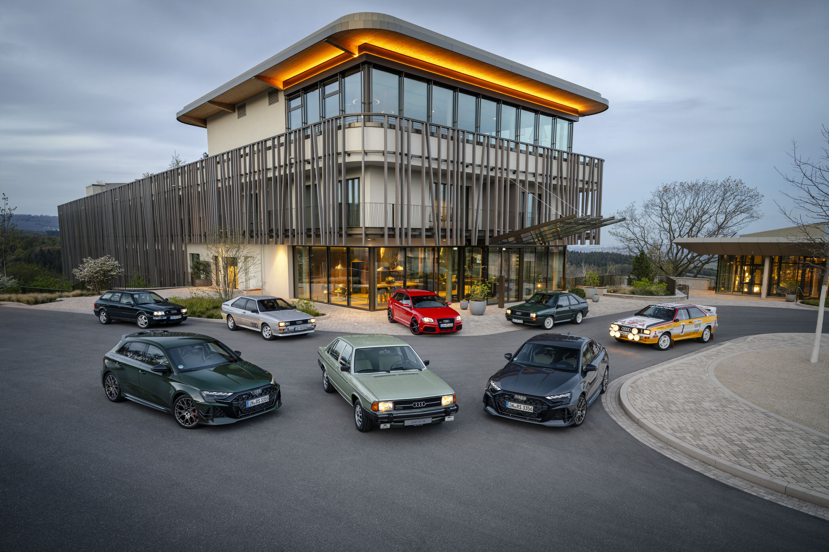 50 Jahre Fünfzylinder - Mehrere Audi Modelle unterschiedlicher Generationen mit Fünfzylindermotor sind halbkreisförmig vor einem modernen Glasgebäude angeordnet.