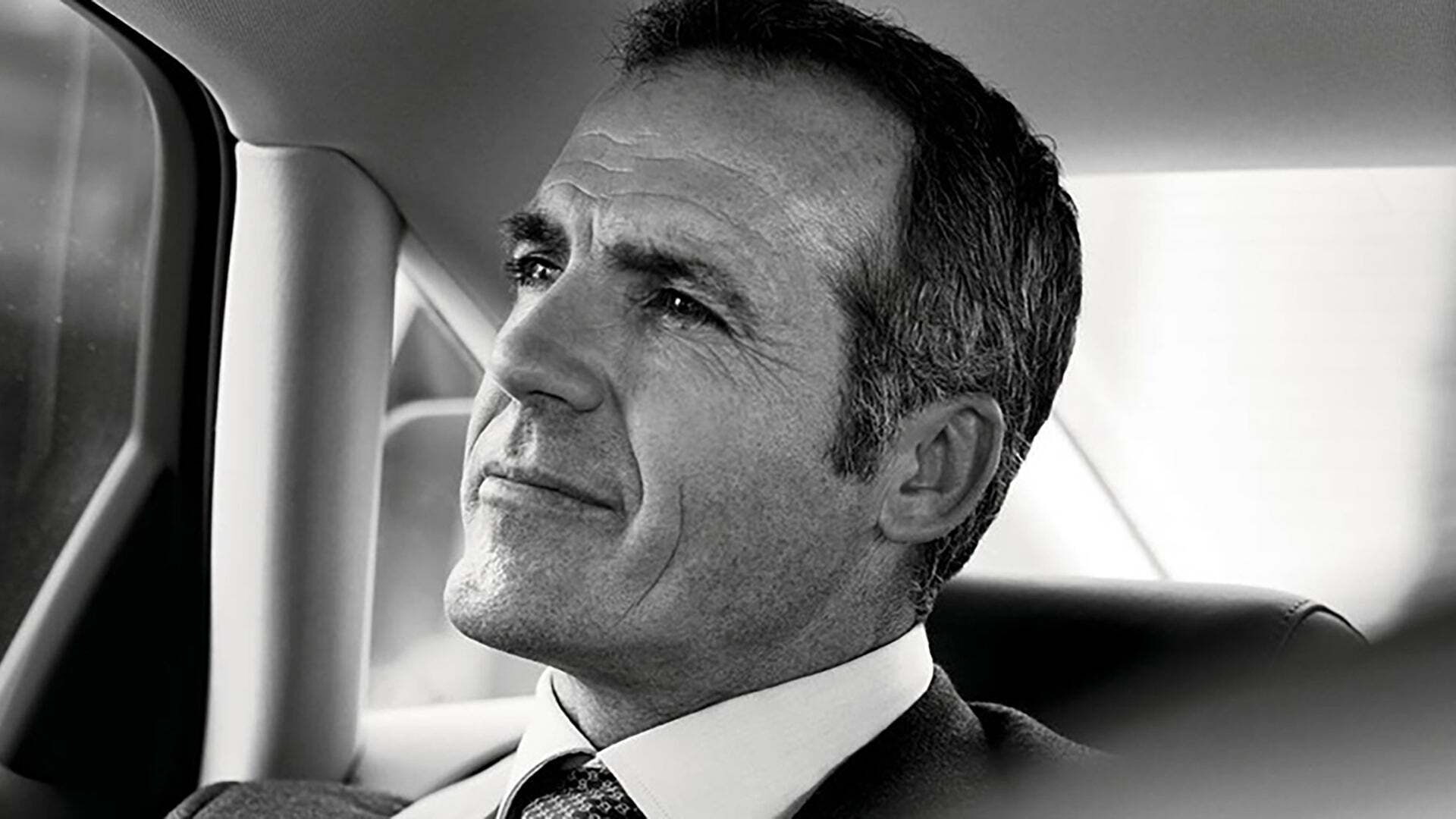 A man in a suit sits in a car, looking thoughtfully out of the window and smiling slightly.