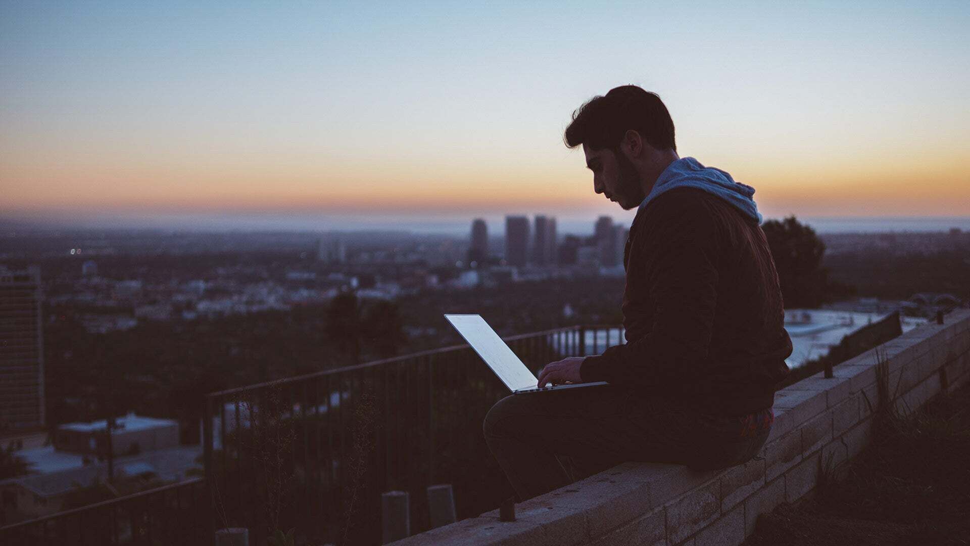 A man sits on a wall at sunrise with his laptop on his lap