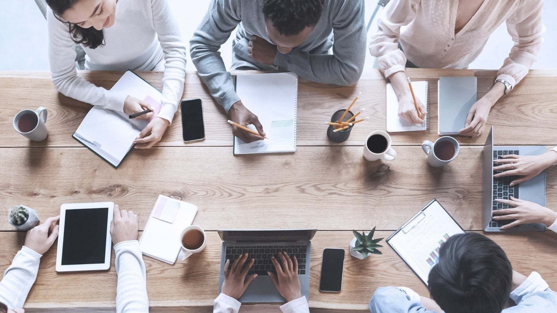 Mehrere Personen sitzen an einem Tisch und arbeiten gemeinsam. Auf dem Tisch sind Laptops, Tablets, Notizen und Kaffeetassen zu sehen.