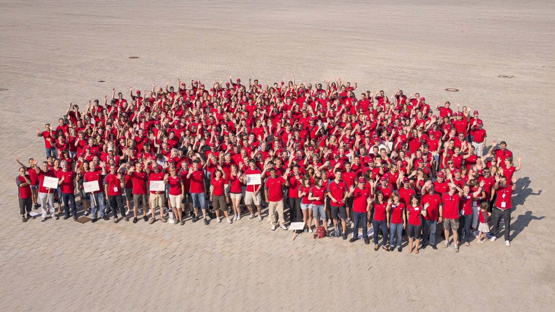 Aerial view of a large group of Audi employees wearing red shirts. They are standing in front of the company headquarters and waving at the camera.