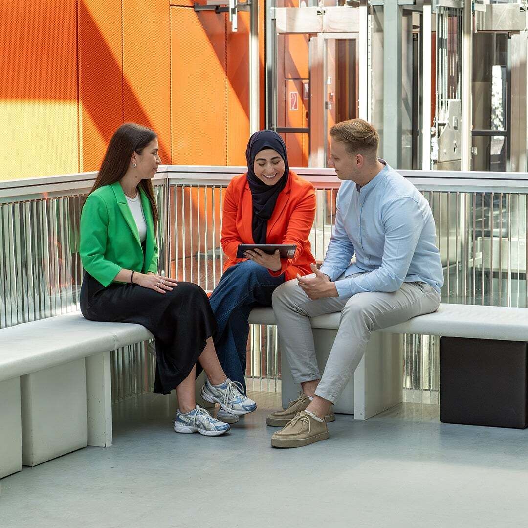 Drei Studenten sitzen auf einer Bank und schauen auf ein Tablet, welches die Mittlere trägt