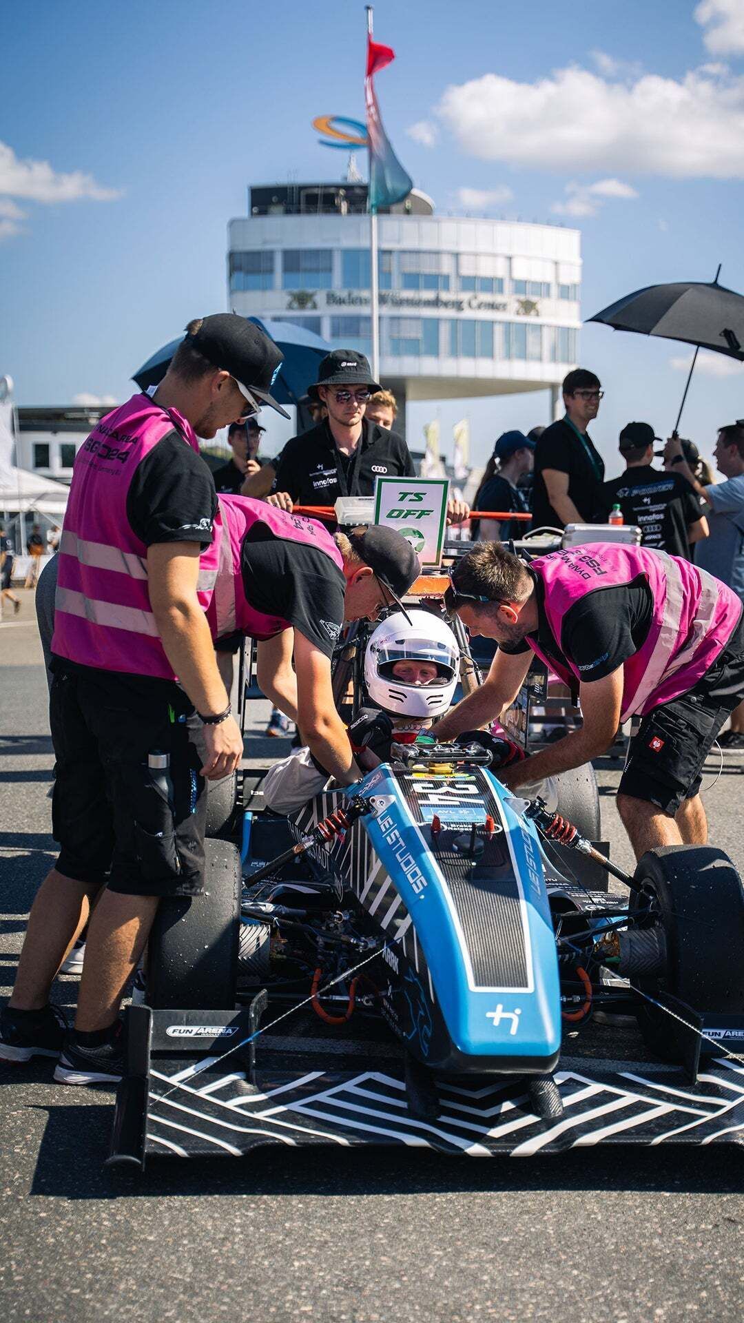 A driver sits in a racing car wearing a helmet and other people stand around the car and work on it