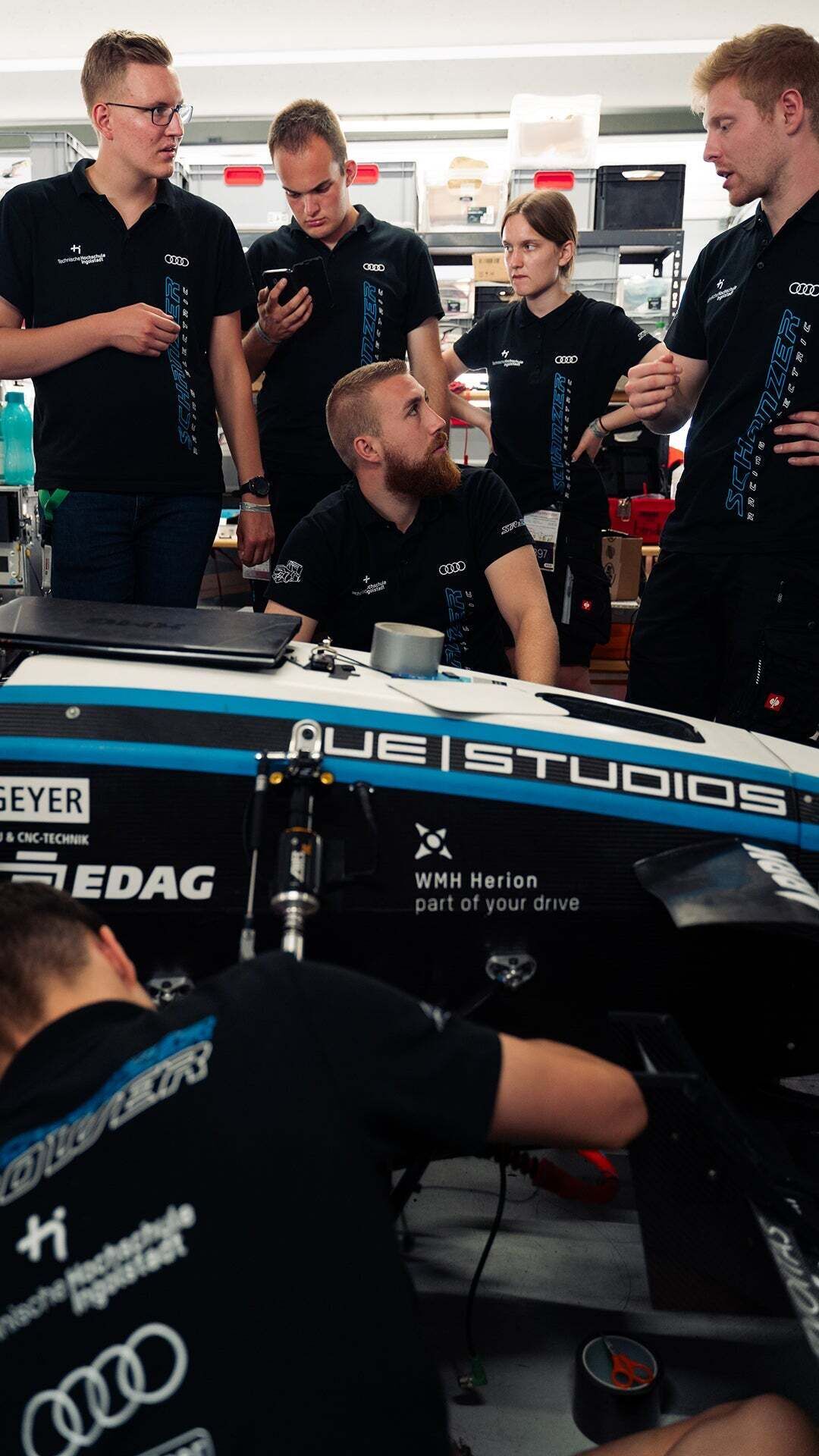 Formula Student - Five people stand behind a racing car and look at each other while a sixth person works on the car.