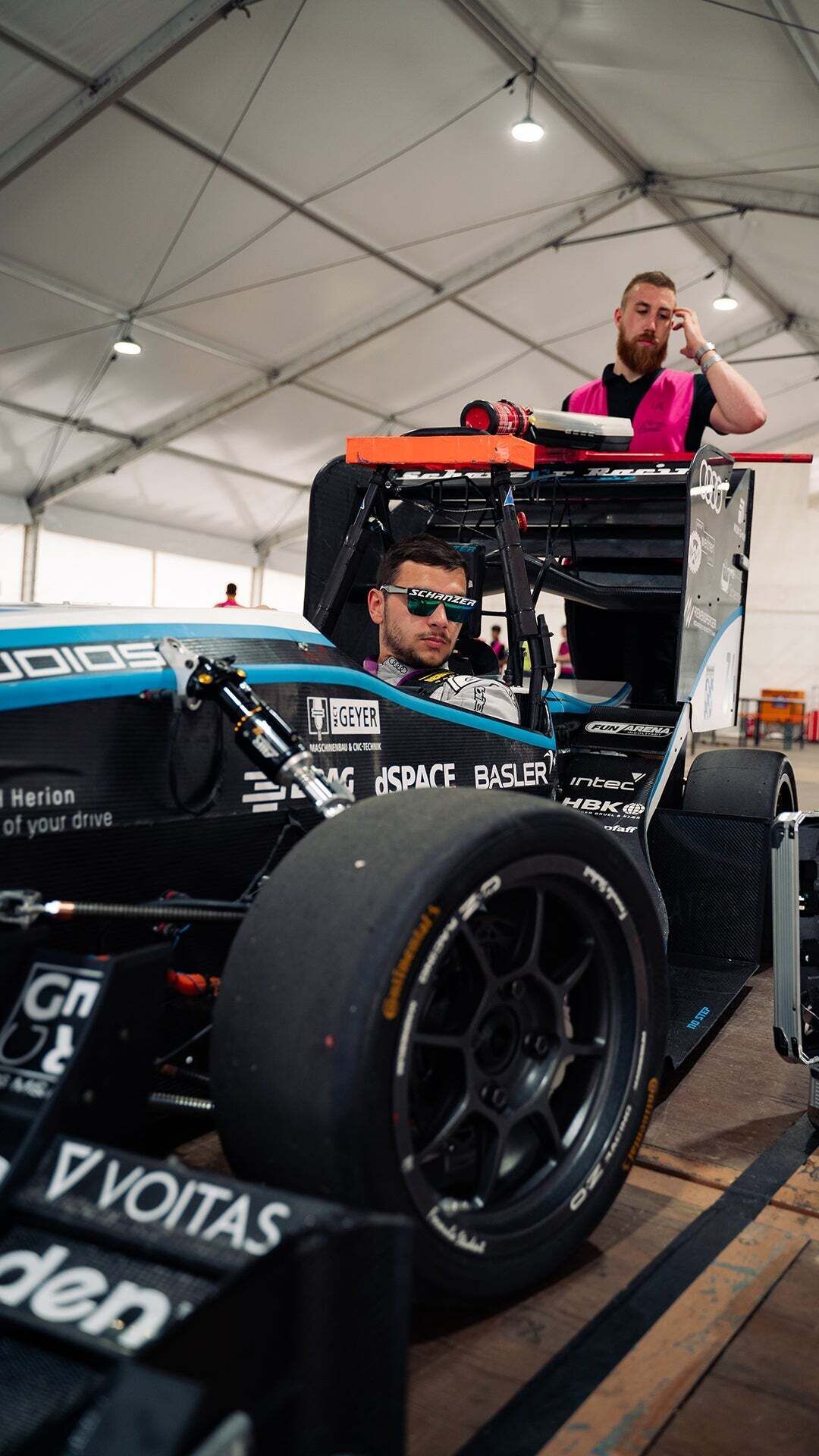 Formula Student - One person sits in a racing car with sunglasses on and another person stands behind the car