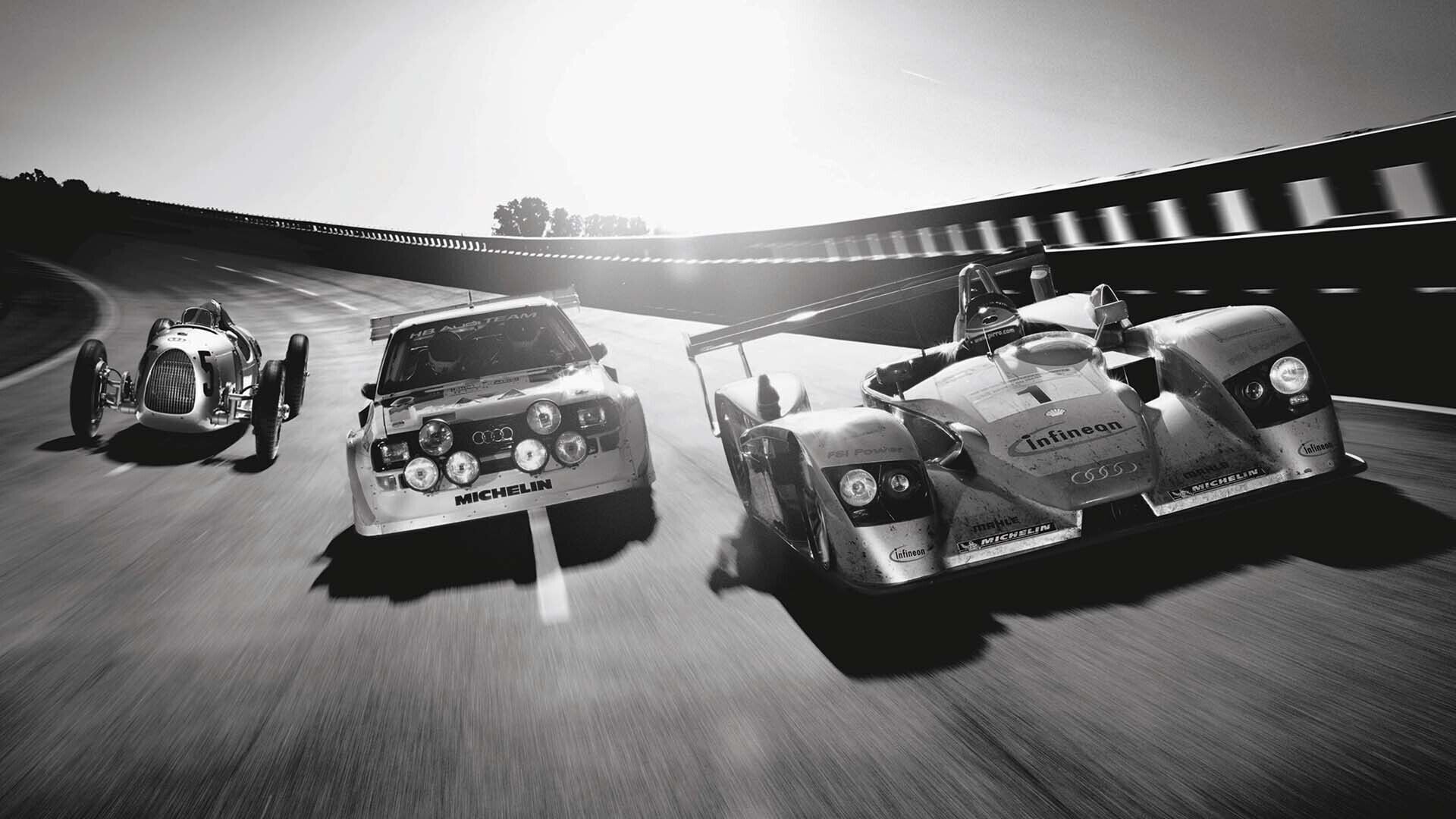 Das Foto zeigt drei historische Audi Rennwagen nebeneinander auf einer abschüssigen Rennstrecke.