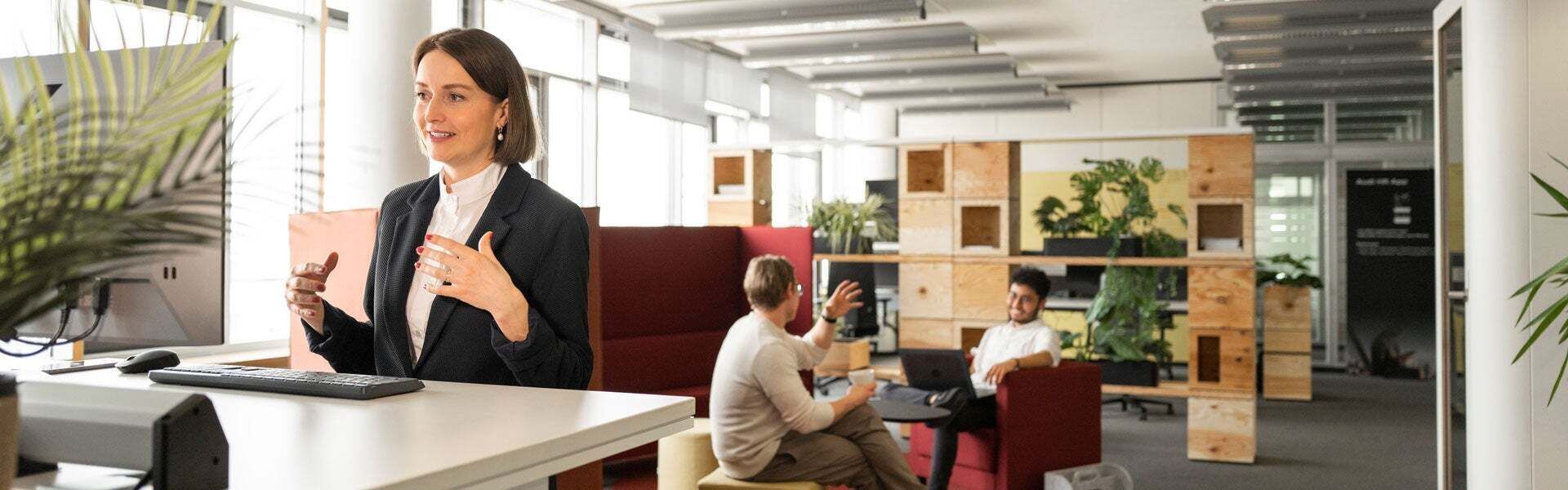 Employee is standing at her desk and colleagues are chatting in the background