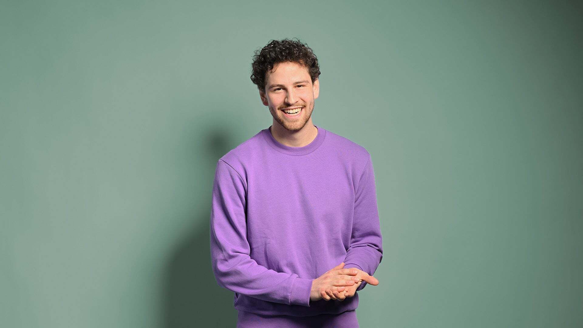 Fabian is standing in front of a wall, wearing a purple sweater, and laughing past the camera.