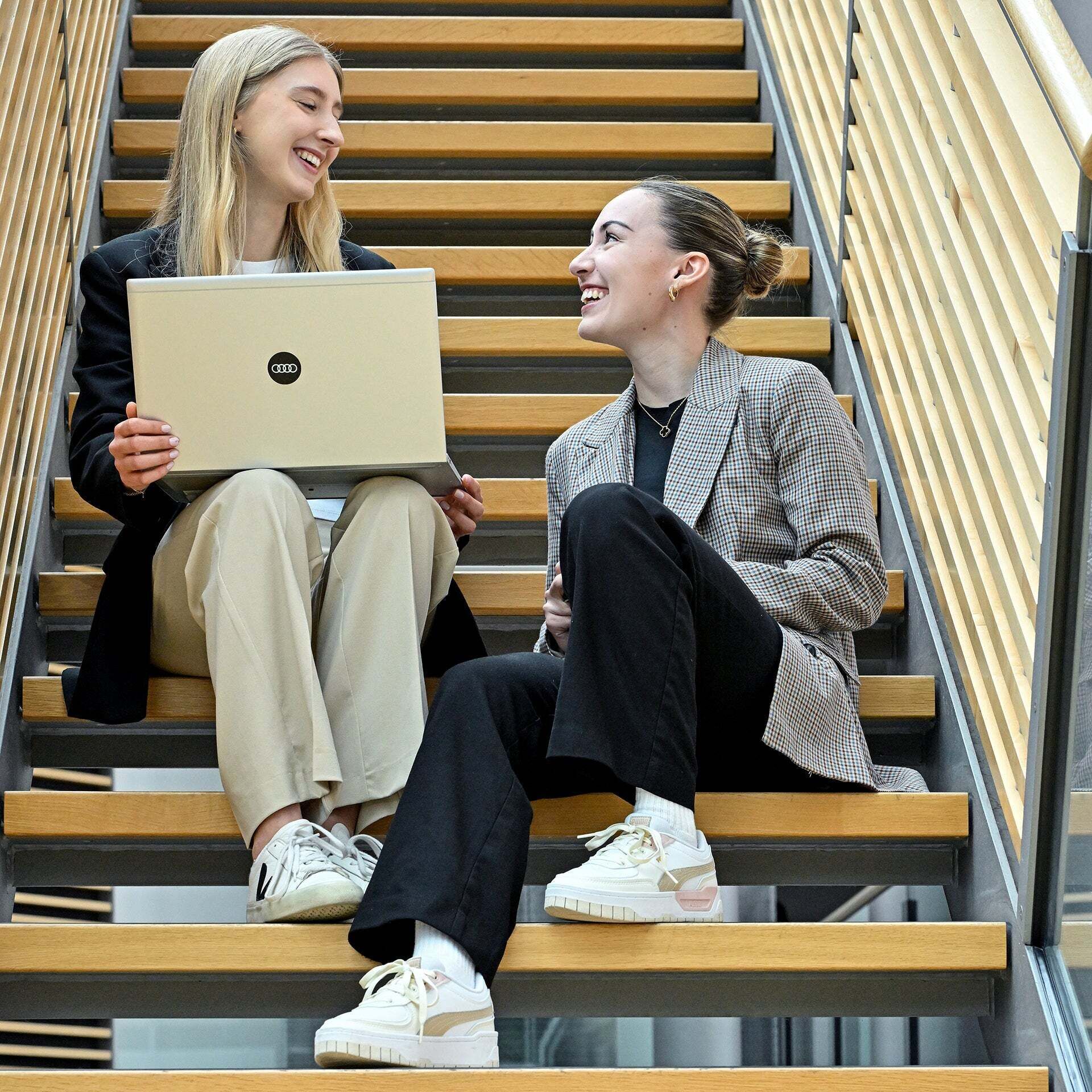 Two employees are sitting on a staircase with a laptop, smiling at each other.